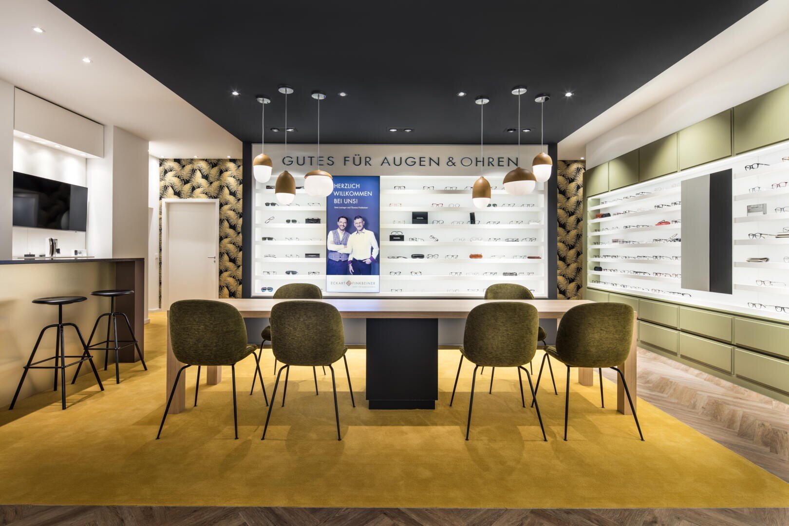 Modernes Optikergeschäft mit Brillenregalen, einem langen Tisch mit grünen Stühlen, Hängelampen und einem Schild in deutscher Sprache mit der Aufschrift "Gutes für Augen & Ohren". Ein digitaler Bildschirm zeigt zwei lächelnde Menschen.