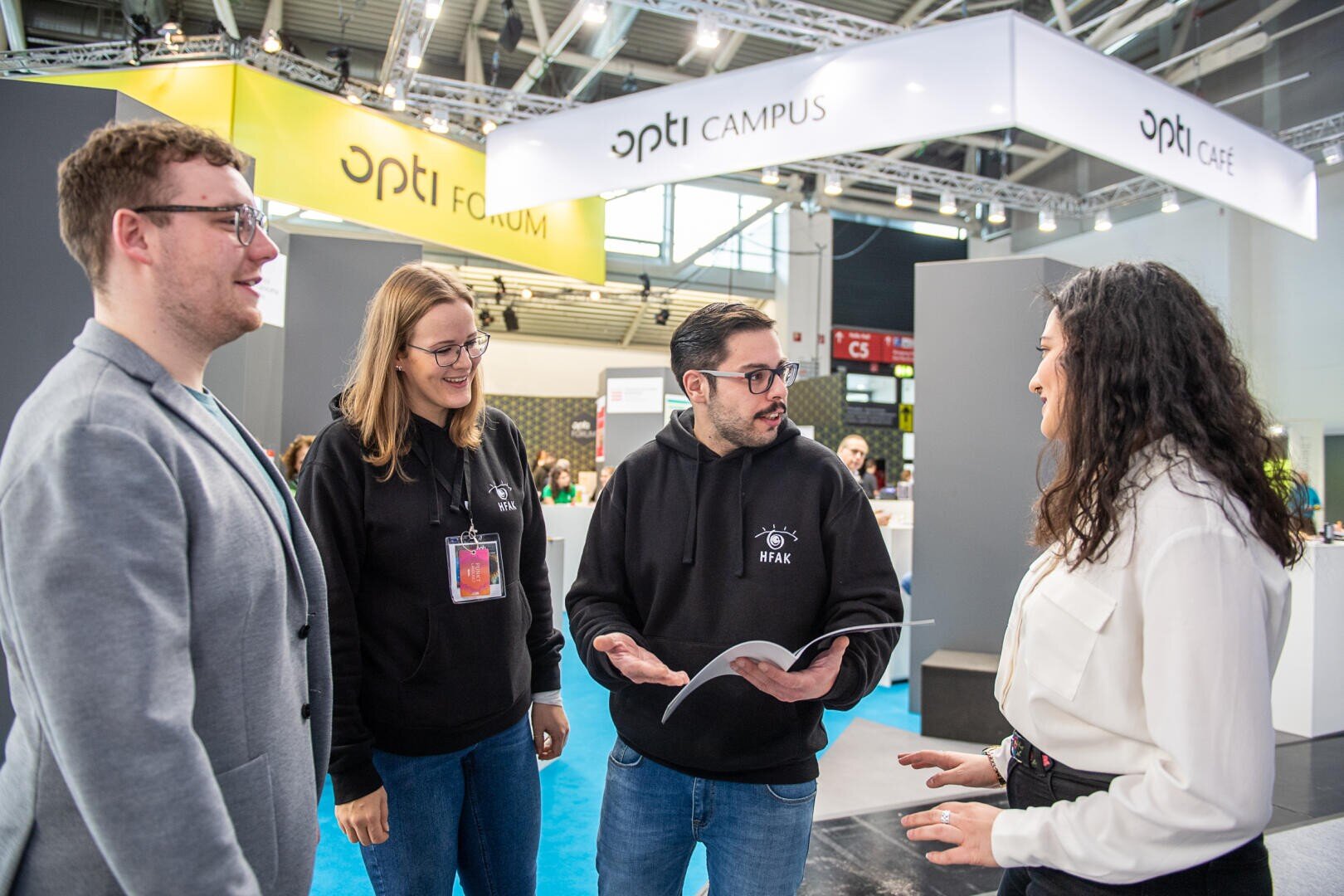 Vier Personen unterhalten sich an einem Messestand. Zwei tragen schwarze Kapuzenpullover, eine eine graue Jacke und eine eine weiße Bluse. Sie wirken in ein Gespräch vertieft, eine hält eine aufgeschlagene Broschüre in der Hand. Auf dem Stand steht „opti CAMPUS“.