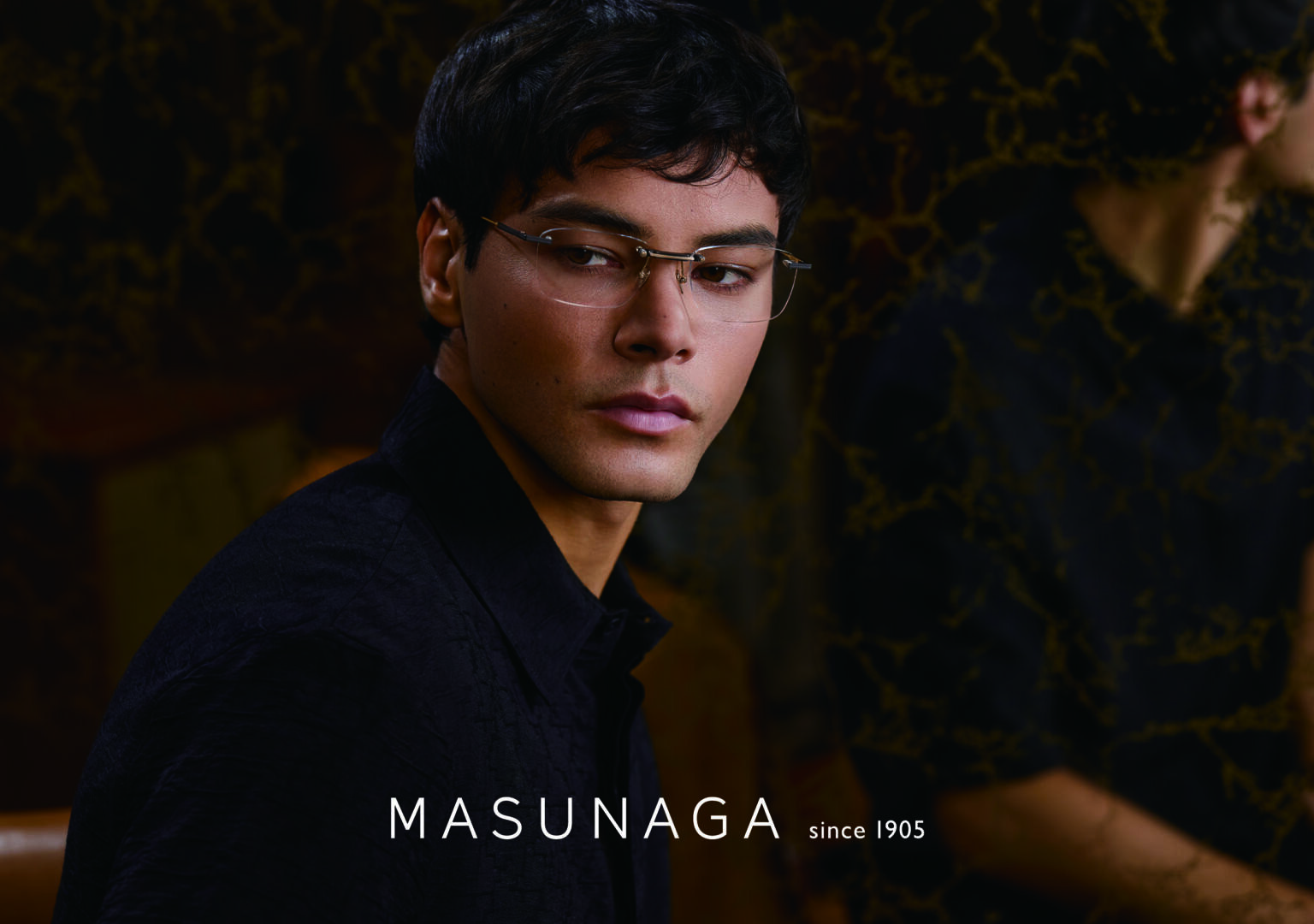 Ein Mann mit kurzen dunklen Haaren und Brille blickt über seine Schulter und trägt ein dunkles Hemd. Der Hintergrund zeigt sein Spiegelbild in einem Spiegel mit Marmormuster. Der Text MASUNAGA since 1905 erscheint am unteren Rand.