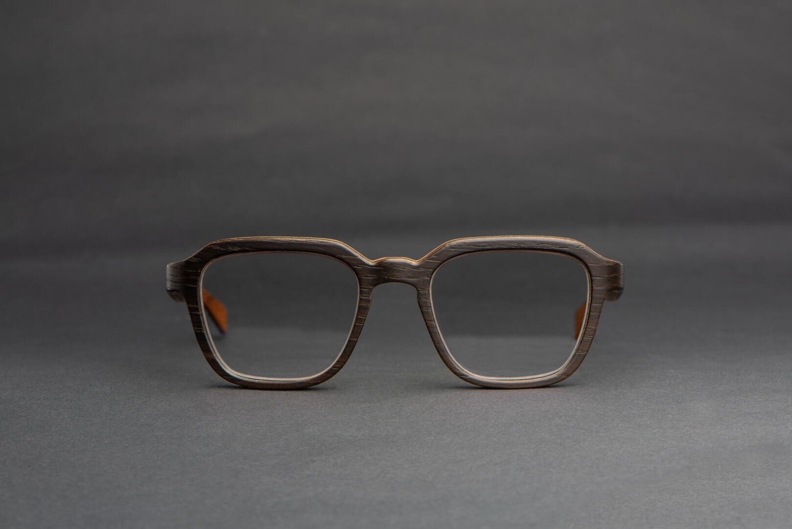 Eine Brille mit einem dunklen, rechteckigen Rahmen mit Holzstruktur ist vor einem schlichten, dunkelgrauen Hintergrund abgebildet.