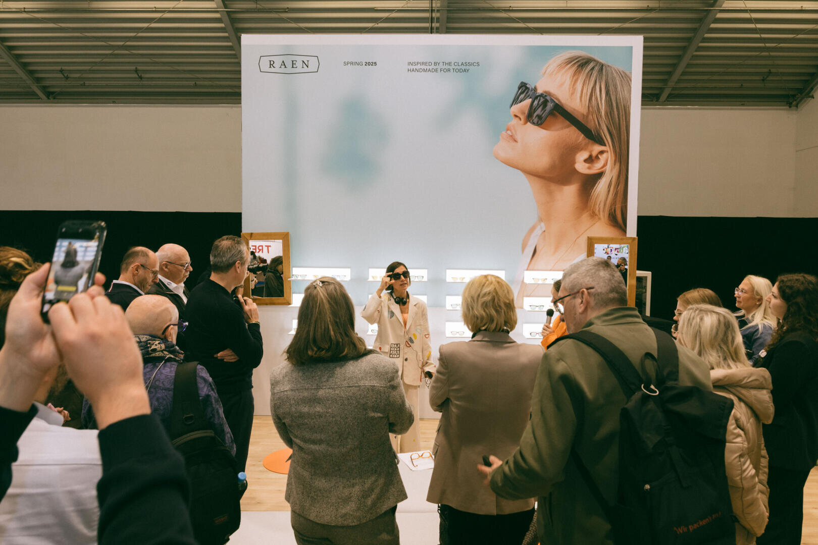 Eine Gruppe von Menschen versammelt sich um eine Frau, die eine Sonnenbrille modelliert, vor einem Ausstellungsstand mit einem großen Poster einer Frau mit Sonnenbrille und der Marke RAEN darüber. Einige Leute machen Fotos.