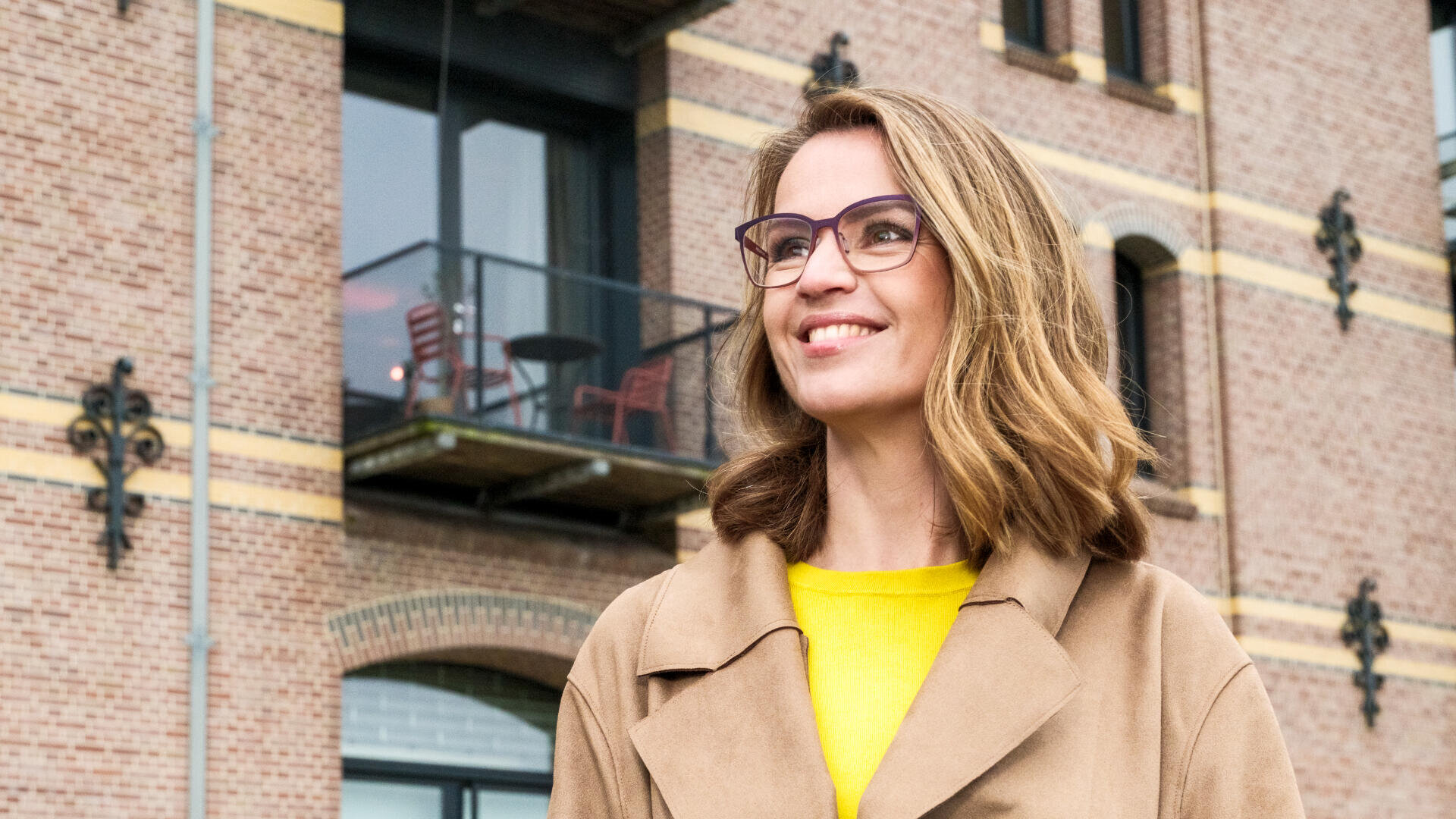A woman with shoulder-length blonde hair and glasses smiles while standing outside in front of a brick building with large windows and a balcony. She is wearing a tan coat over a bright yellow top.