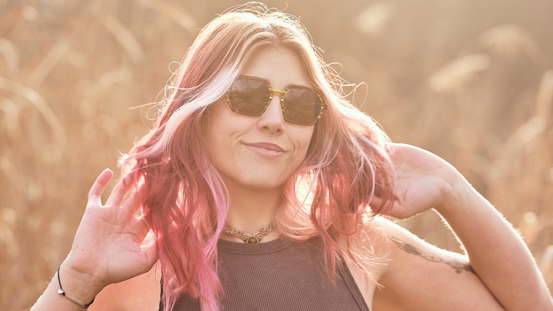 A woman with wavy pink hair and sunglasses smiles outdoors in the sunlight, wearing a sleeveless top and necklace, a hand in her hair and a blurred grassy background.