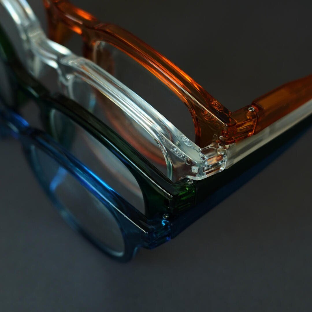 Drei Brillen mit durchsichtigen Rahmen - orange, klar und blau - sind auf einer dunklen Fläche senkrecht gestapelt und aus der Nähe betrachtet.