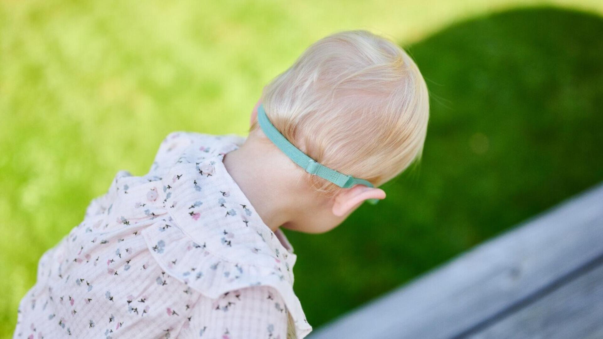 A young child with blonde hair, wearing a light pink patterned shirt and a blue headband, looks down while outdoors on a sunny day with green grass in the background.