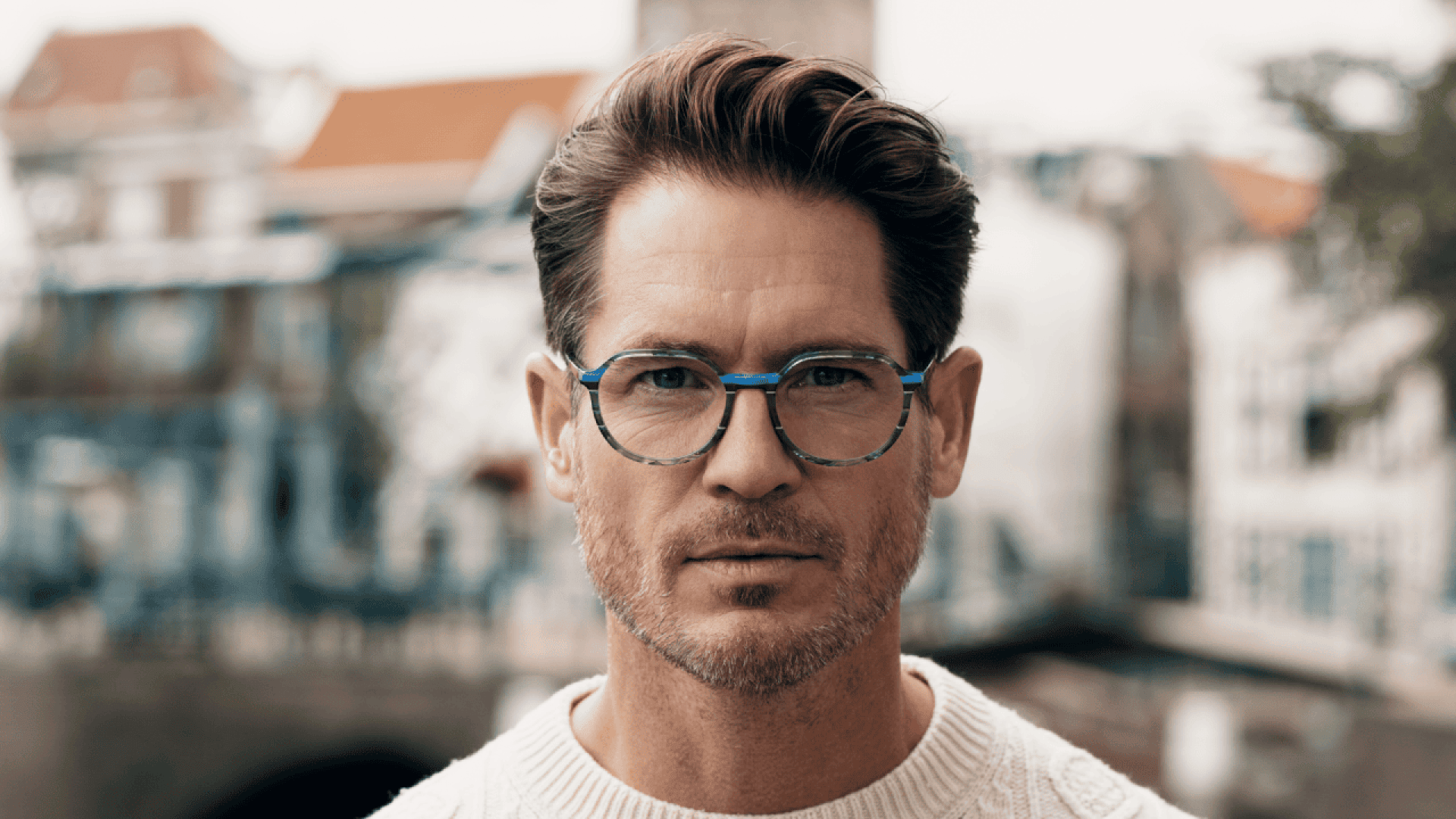 A man with short brown hair, light stubble, and blue-rimmed glasses wears a white sweater and looks directly at the camera. The blurred background shows buildings and a bridge.