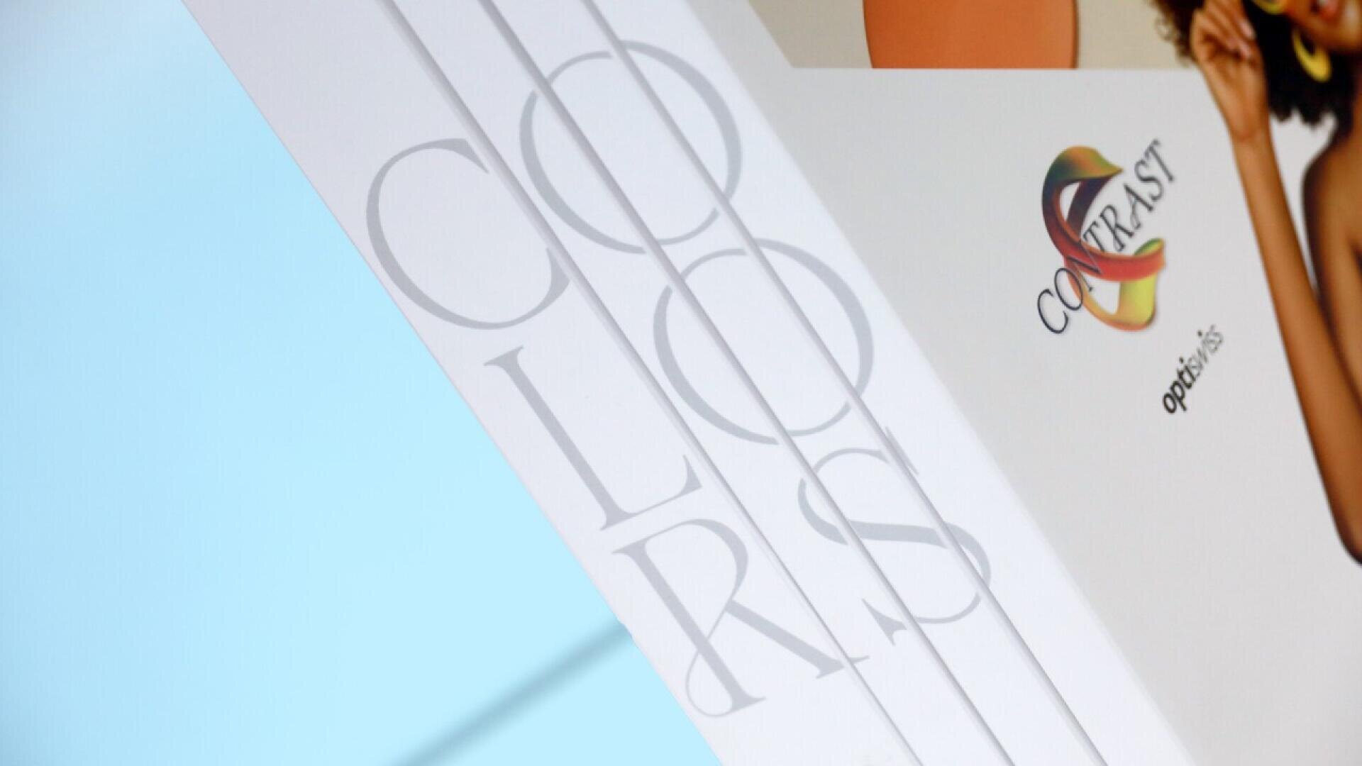A close-up of a product packaging featuring the word “COLORS” in large, light gray letters and a logo with the word “CONTRAST” and “optibliss” on a white background, with a partial image of a person on the right.