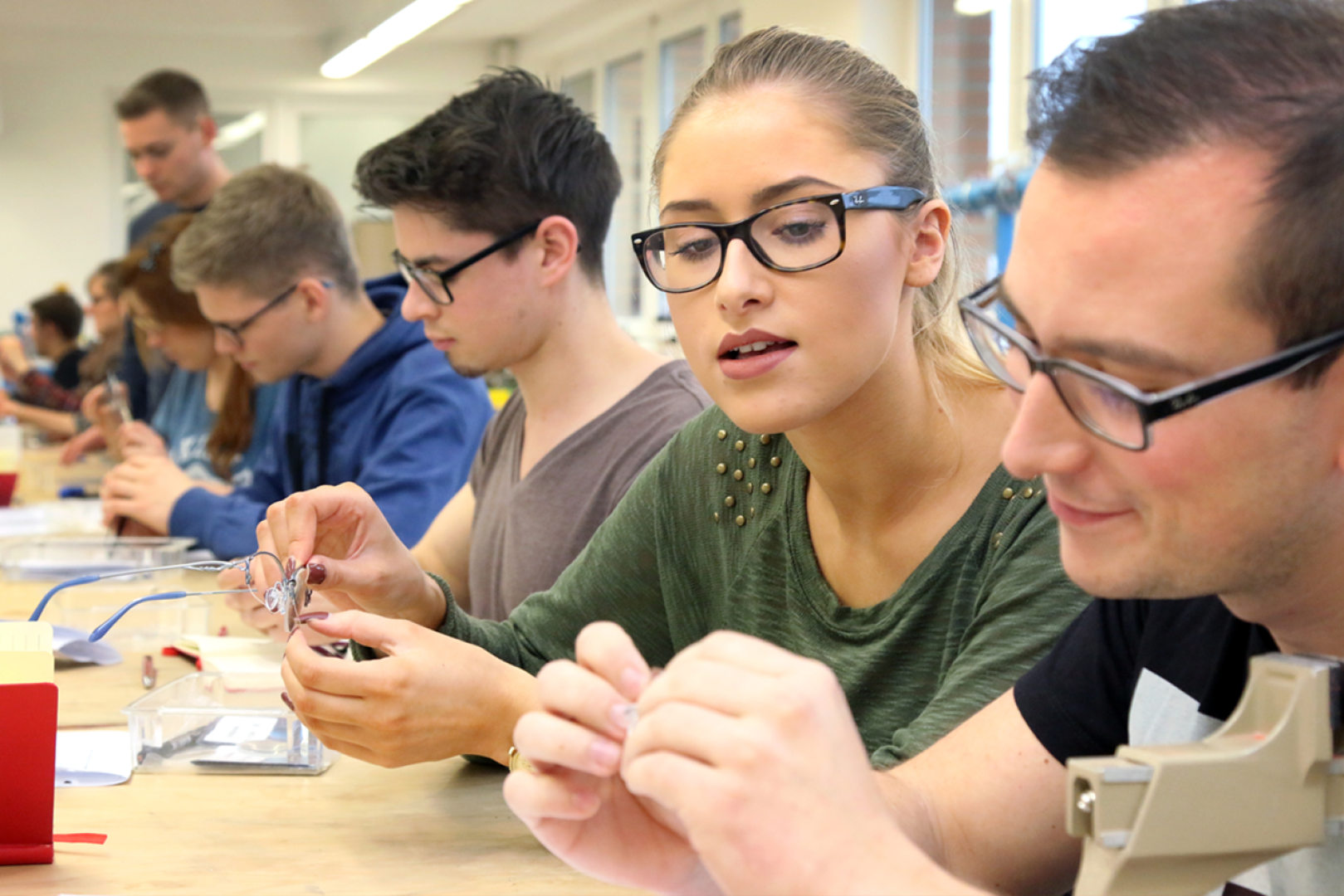 Eine Gruppe junger Erwachsener, die in einem hellen Klassenzimmer an einem Tisch sitzt, arbeitet eng zusammen an praktischen Projekten, hantiert mit kleinen Bauteilen und Werkzeugen, konzentriert und kooperativ.