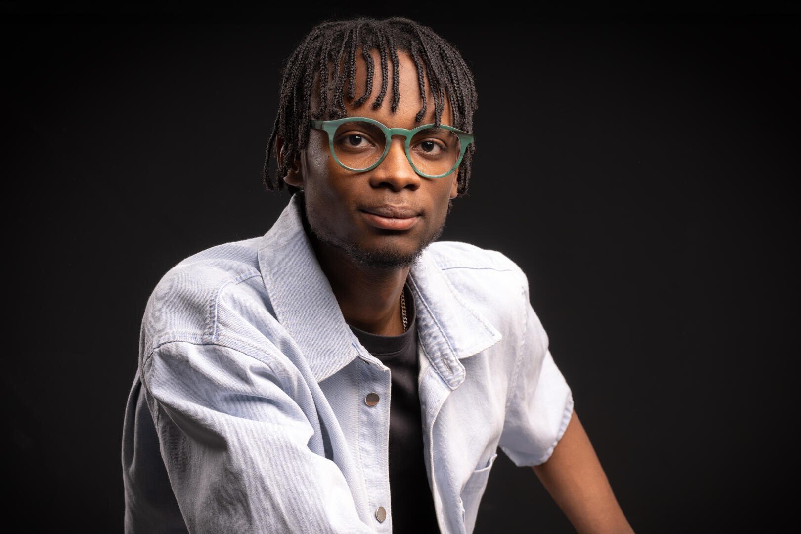 Ein junger Mann mit kurzen Dreadlocks und grün umrandeter Brille trägt eine helle Jeansjacke über einem schwarzen Hemd und posiert vor einem dunklen Hintergrund.