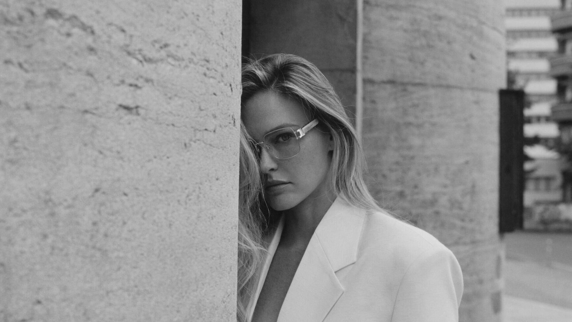 Eine Frau in einem hellen Blazer und mit Brille steht neben einer großen, strukturierten Steinsäule und schaut in die Kamera. Das Bild ist in schwarz-weiß mit einem unscharfen städtischen Hintergrund.