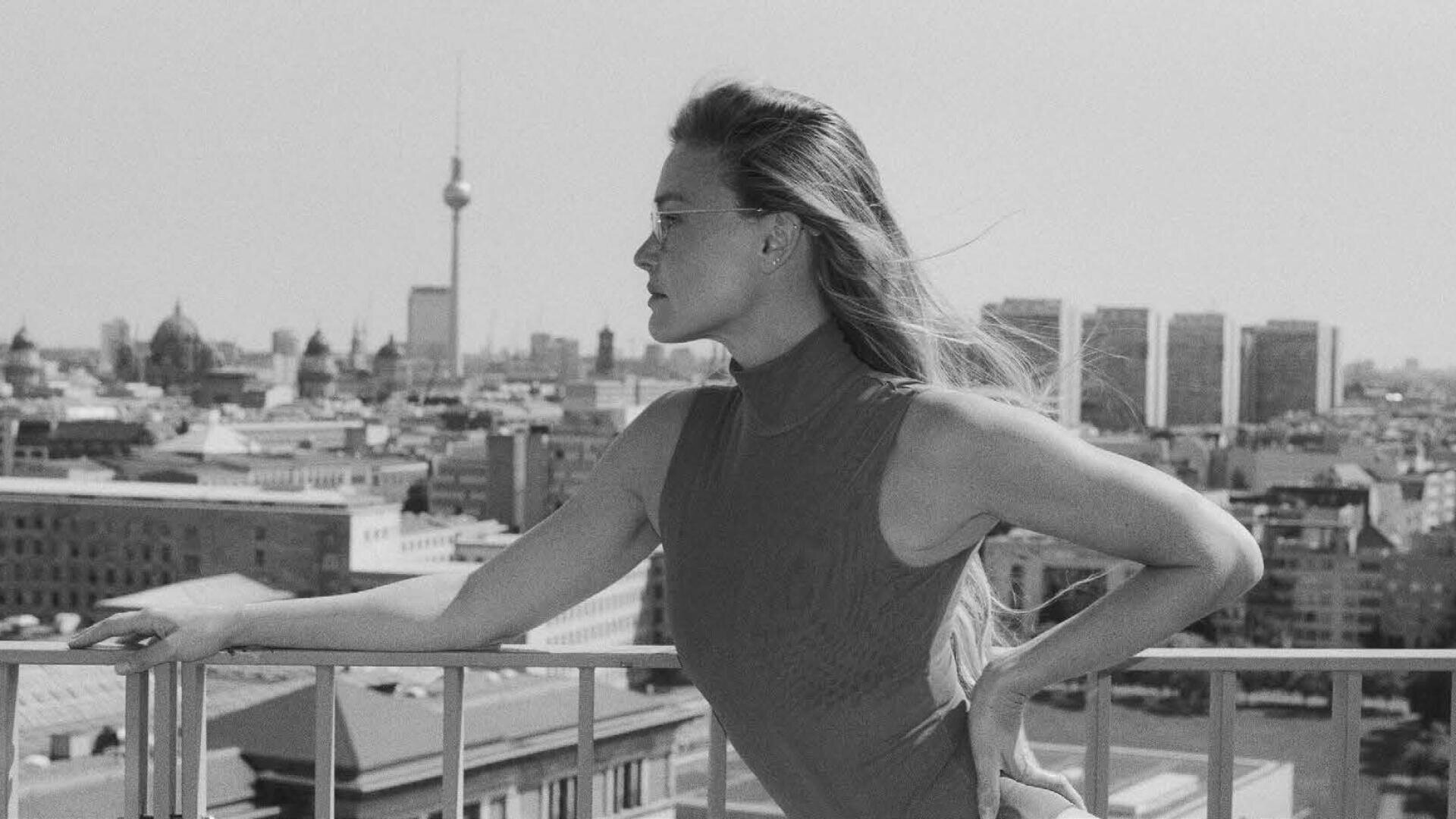 Eine Frau mit langen Haaren und Sonnenbrille lehnt an einem Geländer und blickt auf eine Stadtlandschaft, in der im Hintergrund der Berliner Fernsehturm zu sehen ist. Das Foto ist in schwarz-weiß gehalten.