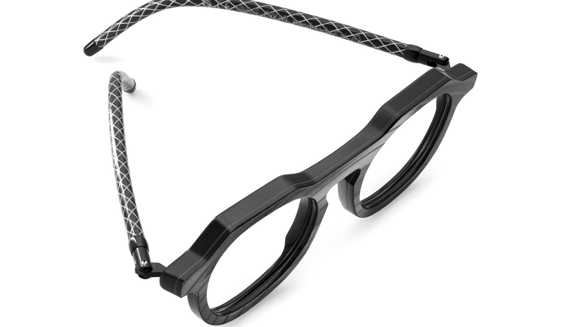 A pair of black round eyeglasses with thick frames and flexible, patterned temples featuring a diamond-like design in white on a gray background. The glasses are positioned at an angle on a white surface.