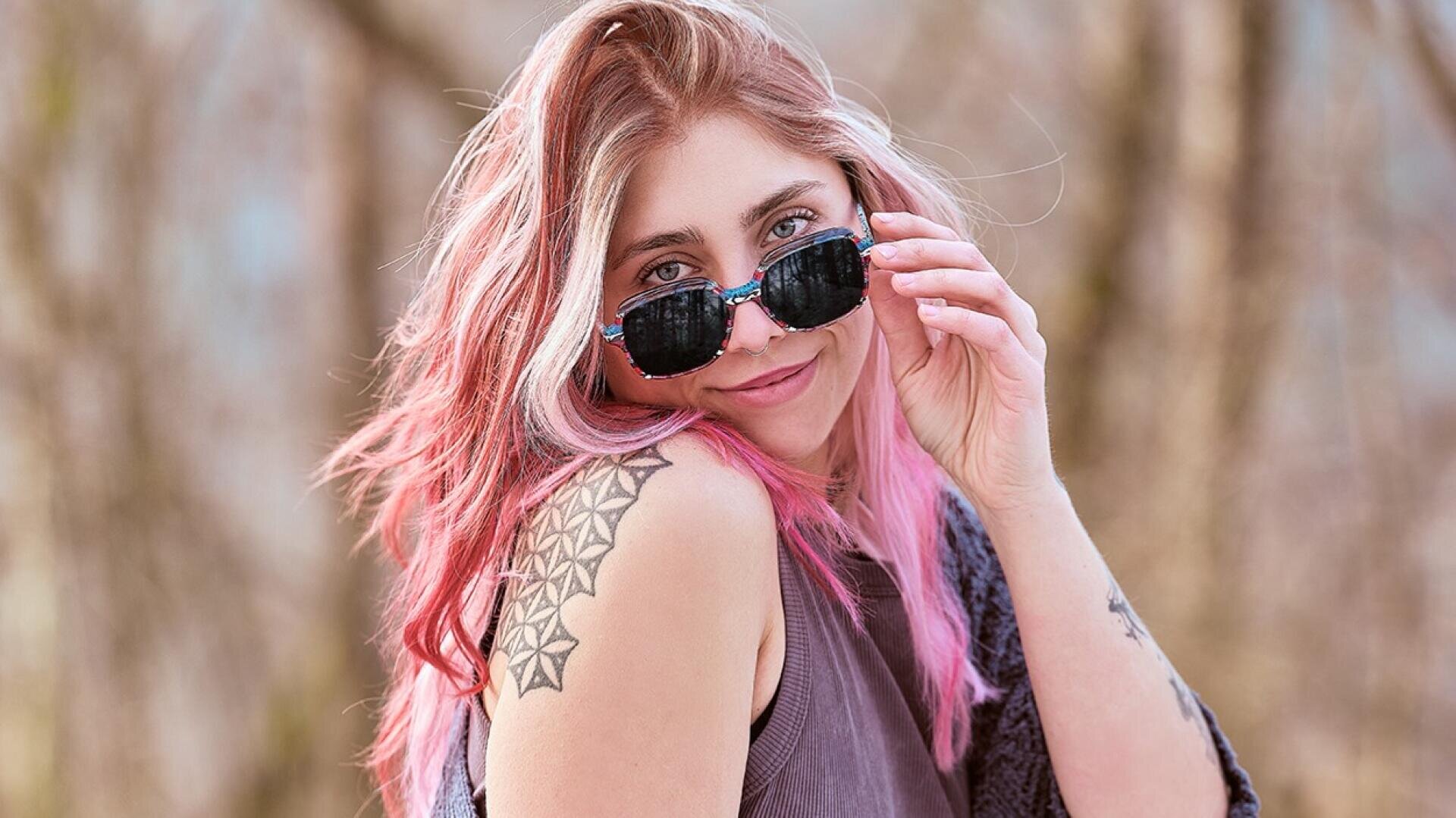 A young woman with long pink hair and a geometric tattoo on her shoulder smiles while holding her sunglasses. She wears a sleeveless top and stands outdoors in front of a blurred natural background.
