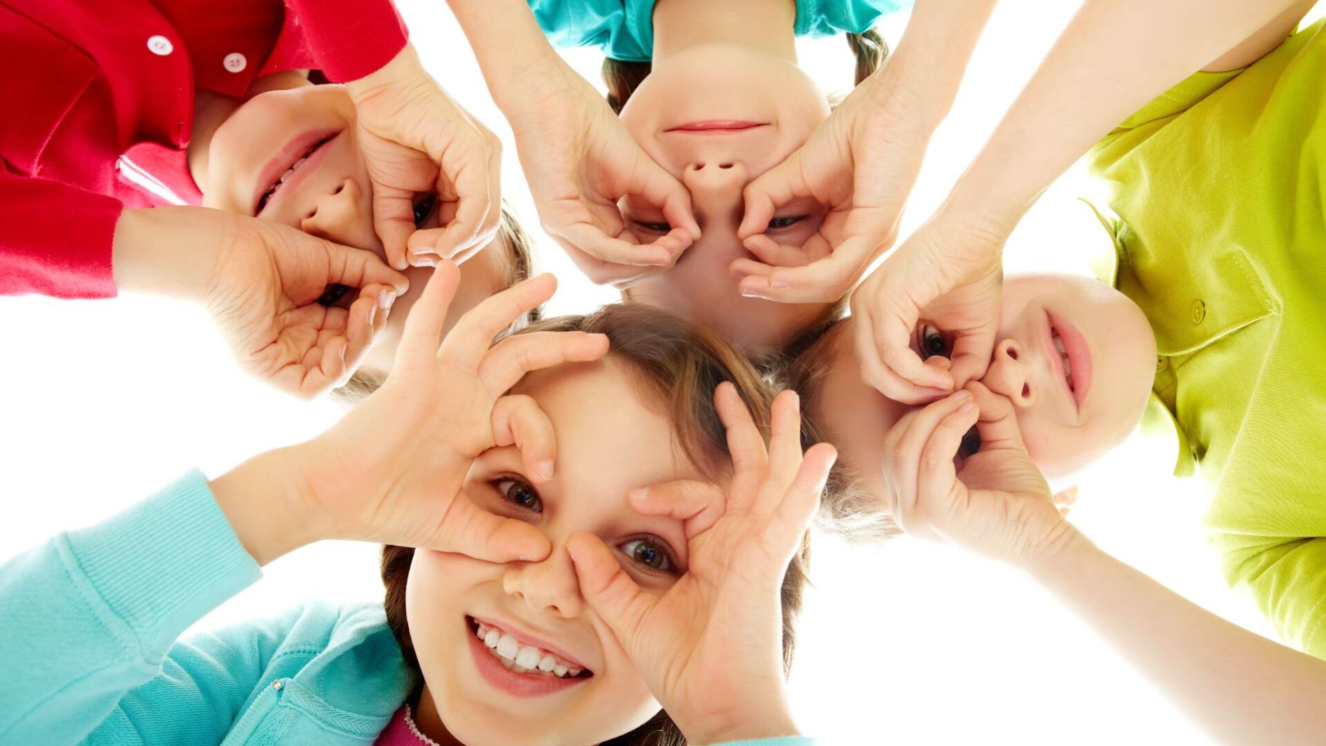 Vier lächelnde Kinder liegen in einem Kreis und blicken in die Kamera. Sie haben sich Handbrillen über die Augen gezogen und tragen bunte Hemden in Rot, Blau, Grün und Türkis. Der Hintergrund ist strahlend weiß.