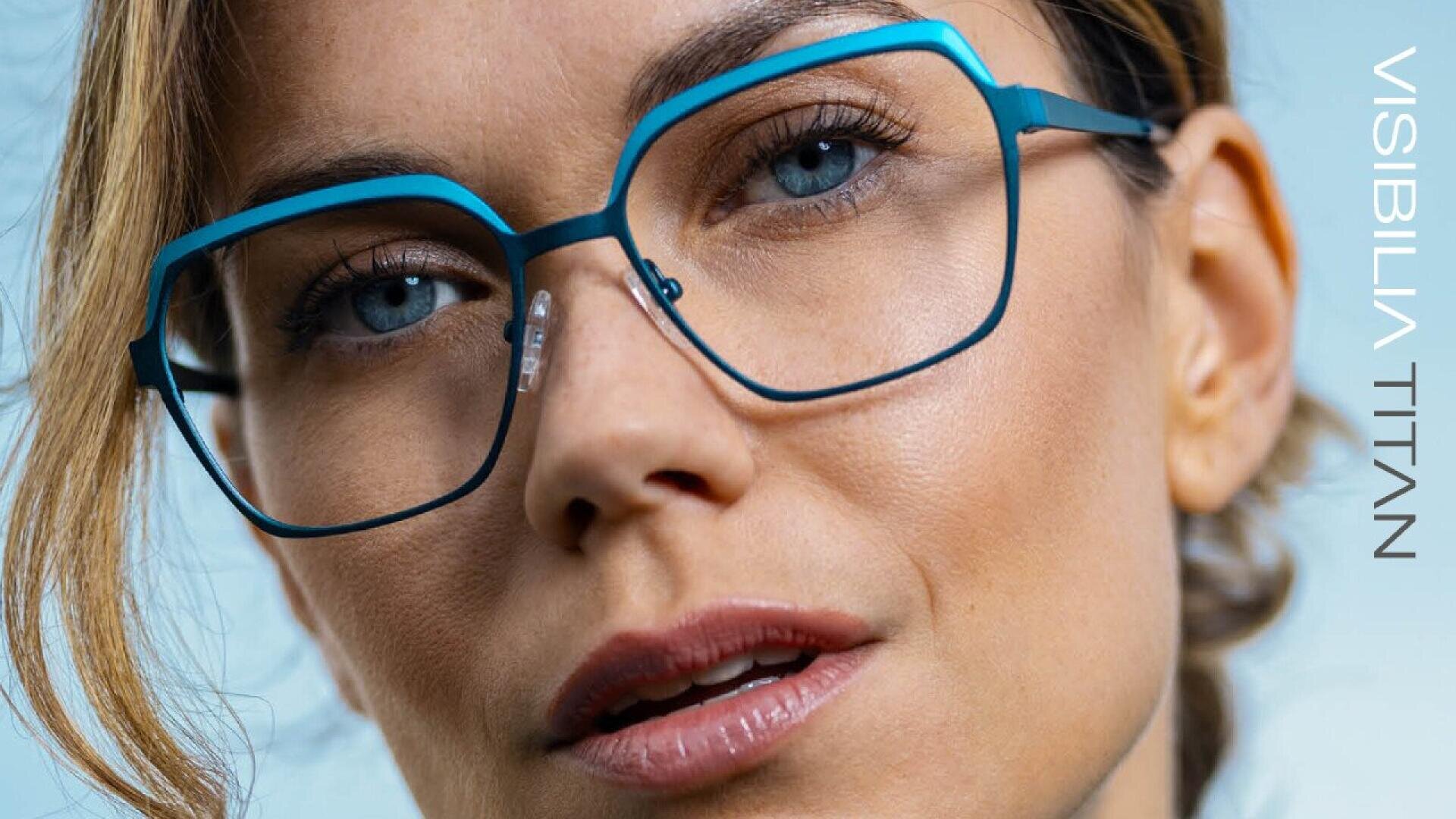 Close-up of a woman wearing stylish, blue-rimmed eyeglasses with a soft expression. Text on the right reads “VISIBILIA TITAN.” The background is a light, neutral shade.