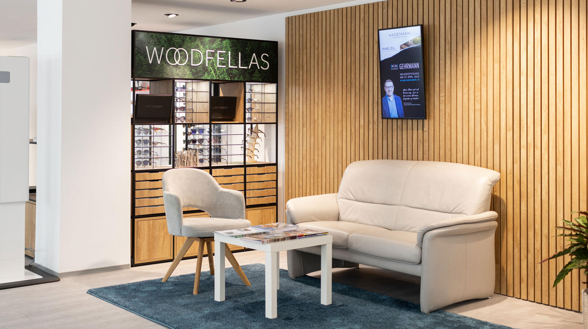 A modern waiting area with a beige sofa, gray chair, small white table with magazines, and a blue rug. Behind, there’s a wooden display of eyeglass frames and a sign reading “WOODFELLAS.” A TV is mounted on a wood-paneled wall.
