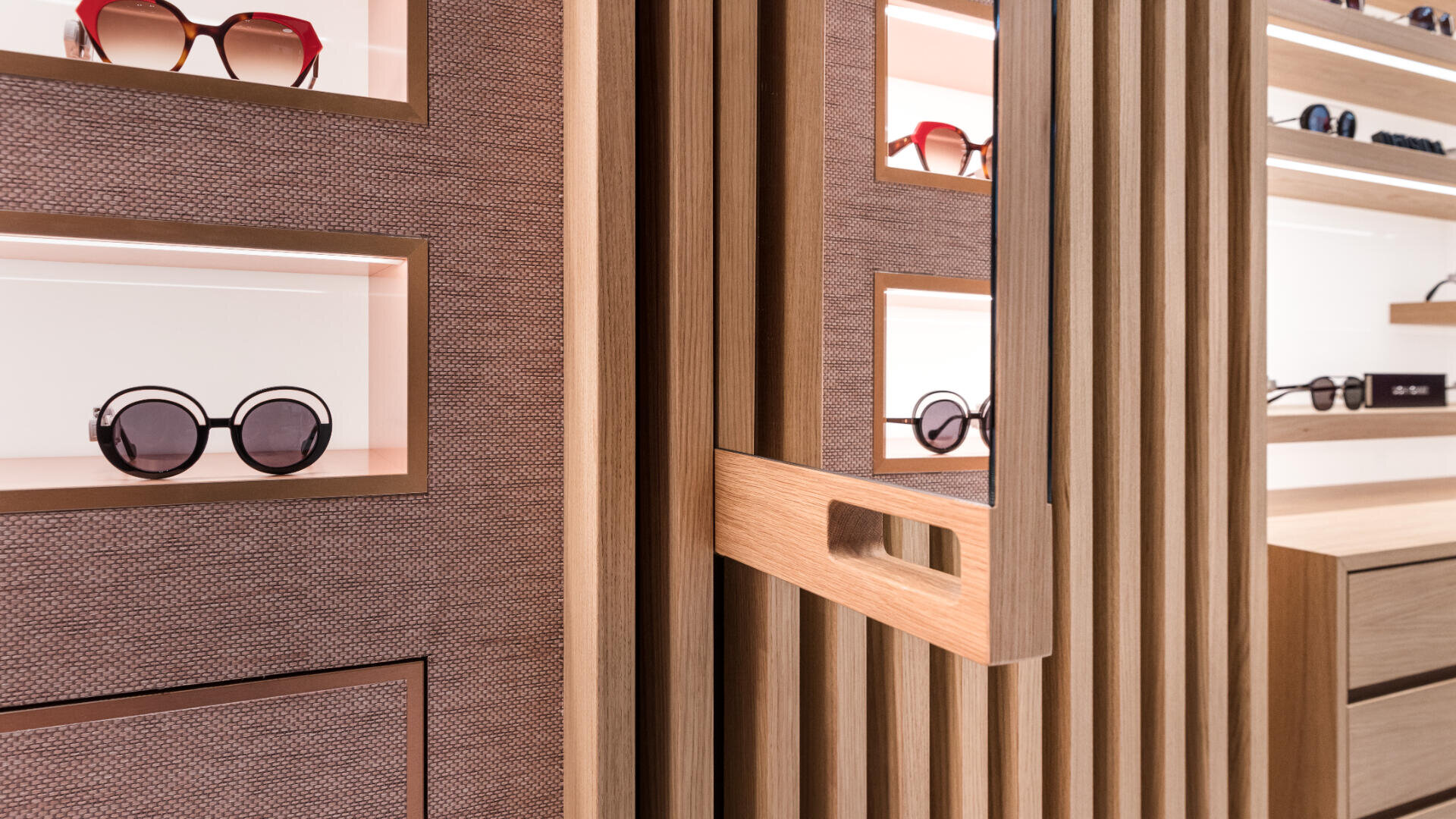 Eine moderne Brillenausstellung präsentiert Sonnenbrillen auf beleuchteten Regalen in strukturierten Wänden mit vertikalen Holzpaneelen und einem Griff im Vordergrund. Die Szene besticht durch eine klare, minimalistische Ästhetik.