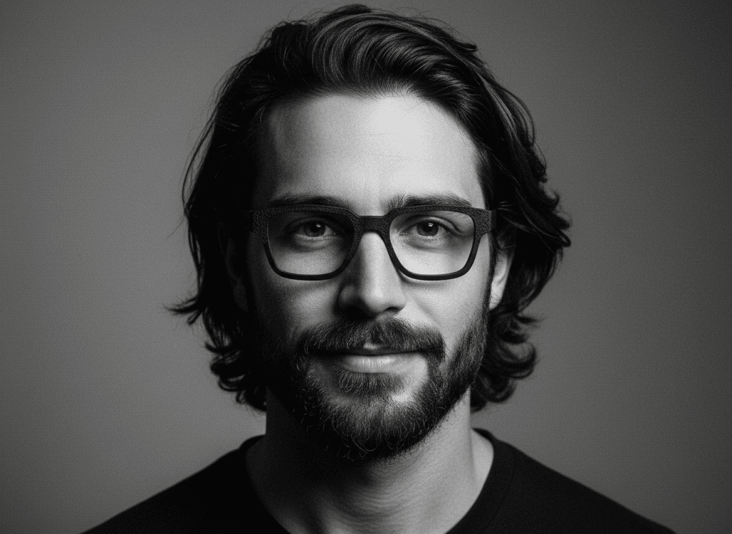 A man with medium-length, wavy hair, a beard and glasses looks directly into the camera and smiles slightly. The black and white portrait has a simple, softly lit background.