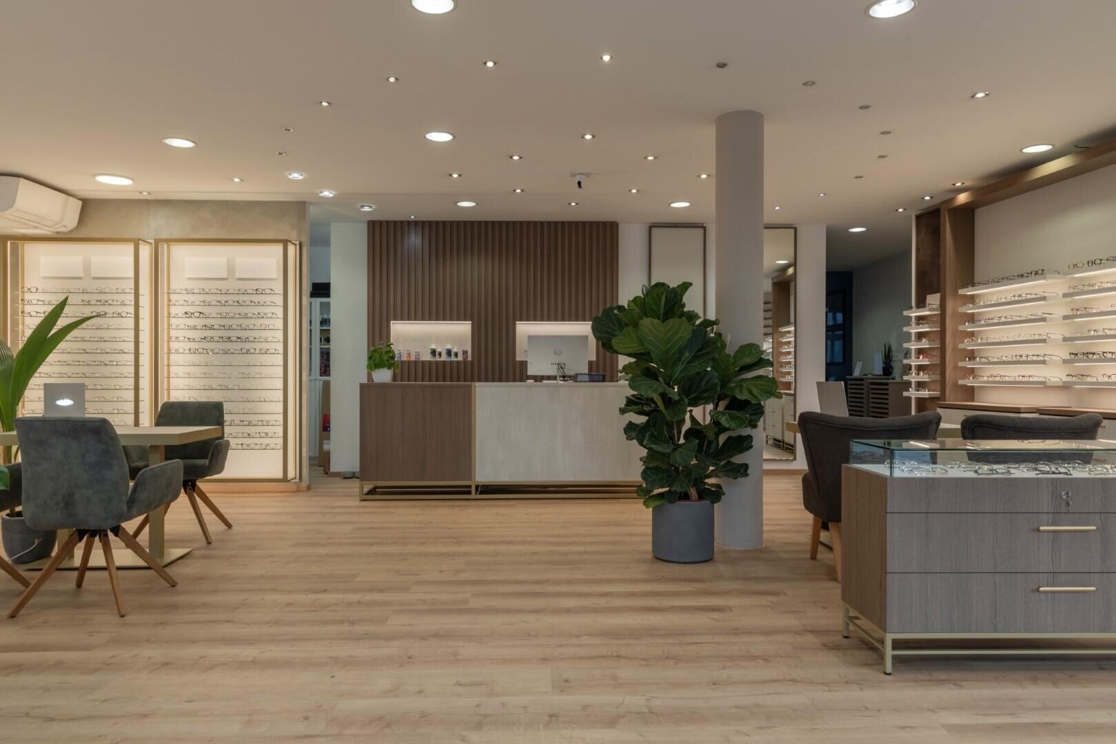 Die Inneneinrichtung eines modernen Optikergeschäfts mit Holzböden, mit Brillen gefüllten Regalen, einem zentralen Check-in-Schalter, grauen Stühlen, Pflanzen und warmer Beleuchtung schafft eine einladende Atmosphäre.