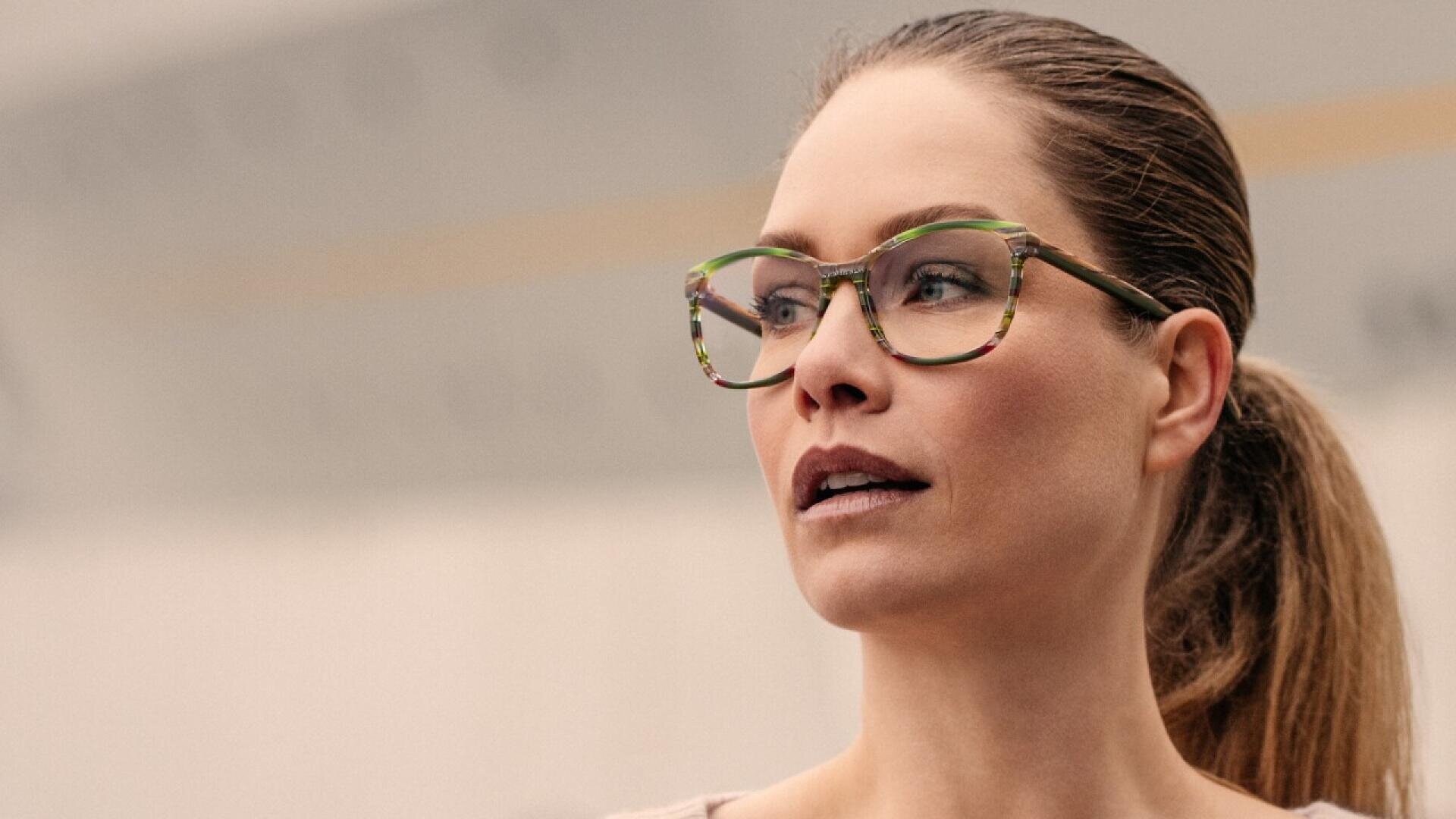 A woman with brown hair tied back wears large, green-rimmed eyeglasses and looks slightly to the side with a neutral expression. The background is softly blurred.