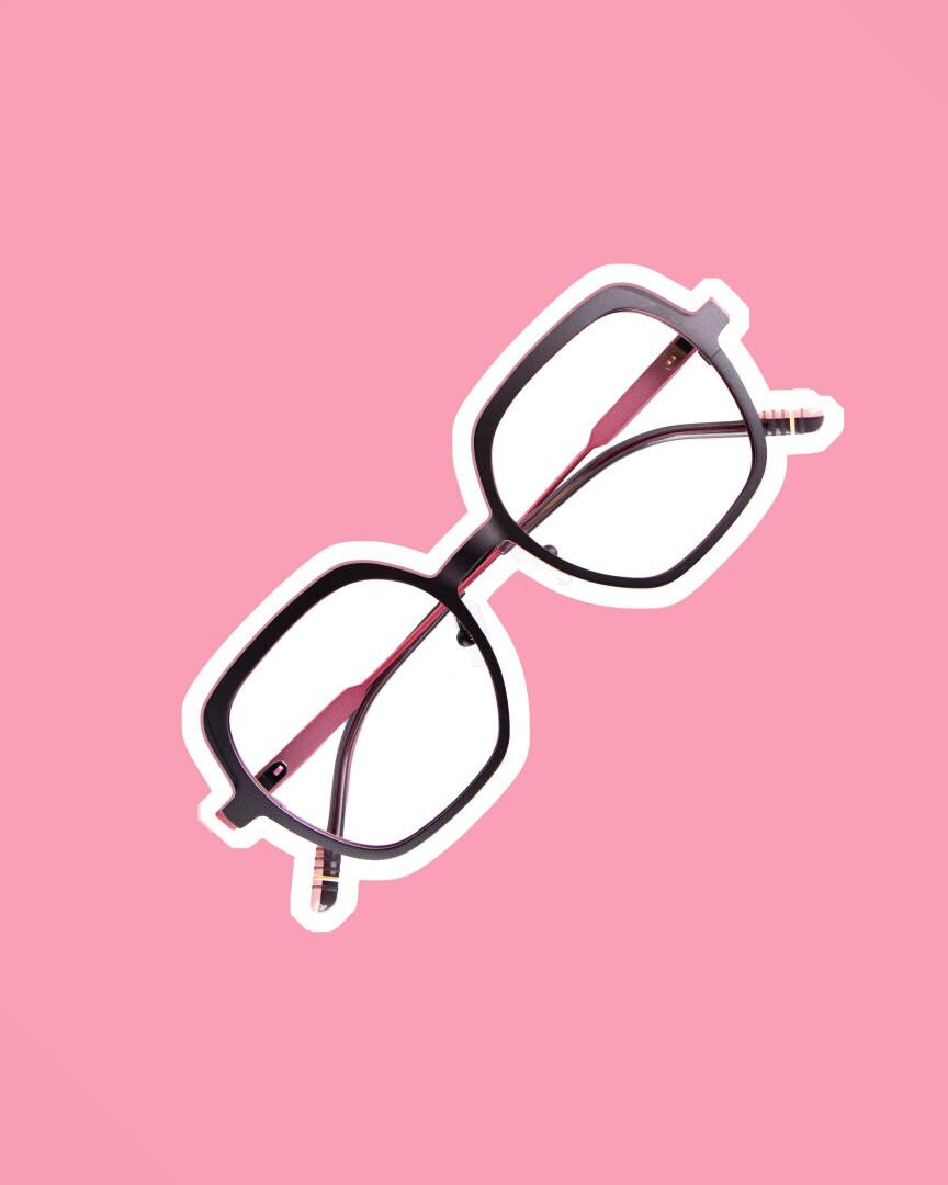 A pair of black, square-framed glasses with clear lenses sits on a pink background. The picture has a white outline around the glasses to emphasize them.