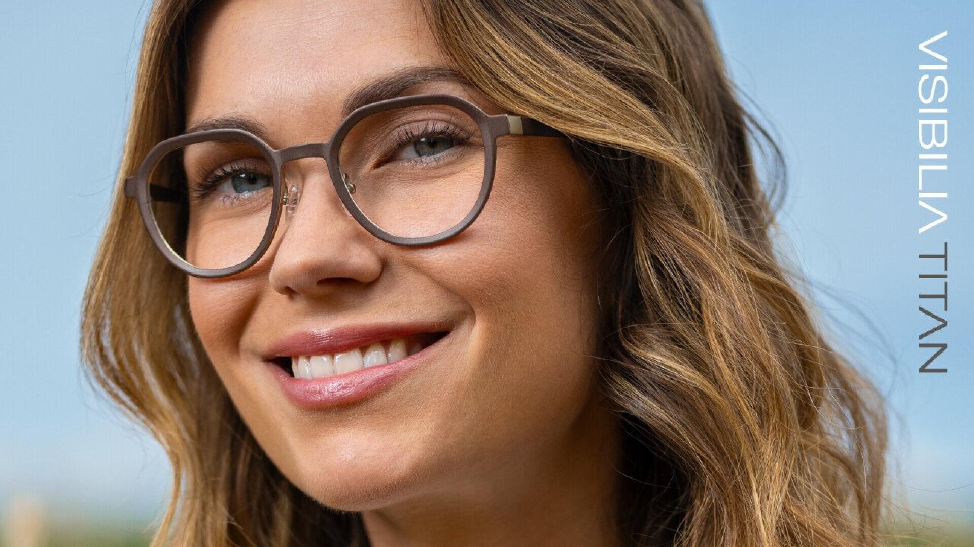A woman with wavy light brown hair smiles while wearing round, brown eyeglasses. The text VISIBILIA TITAN is vertically displayed on the right side of the image. The background is softly blurred.