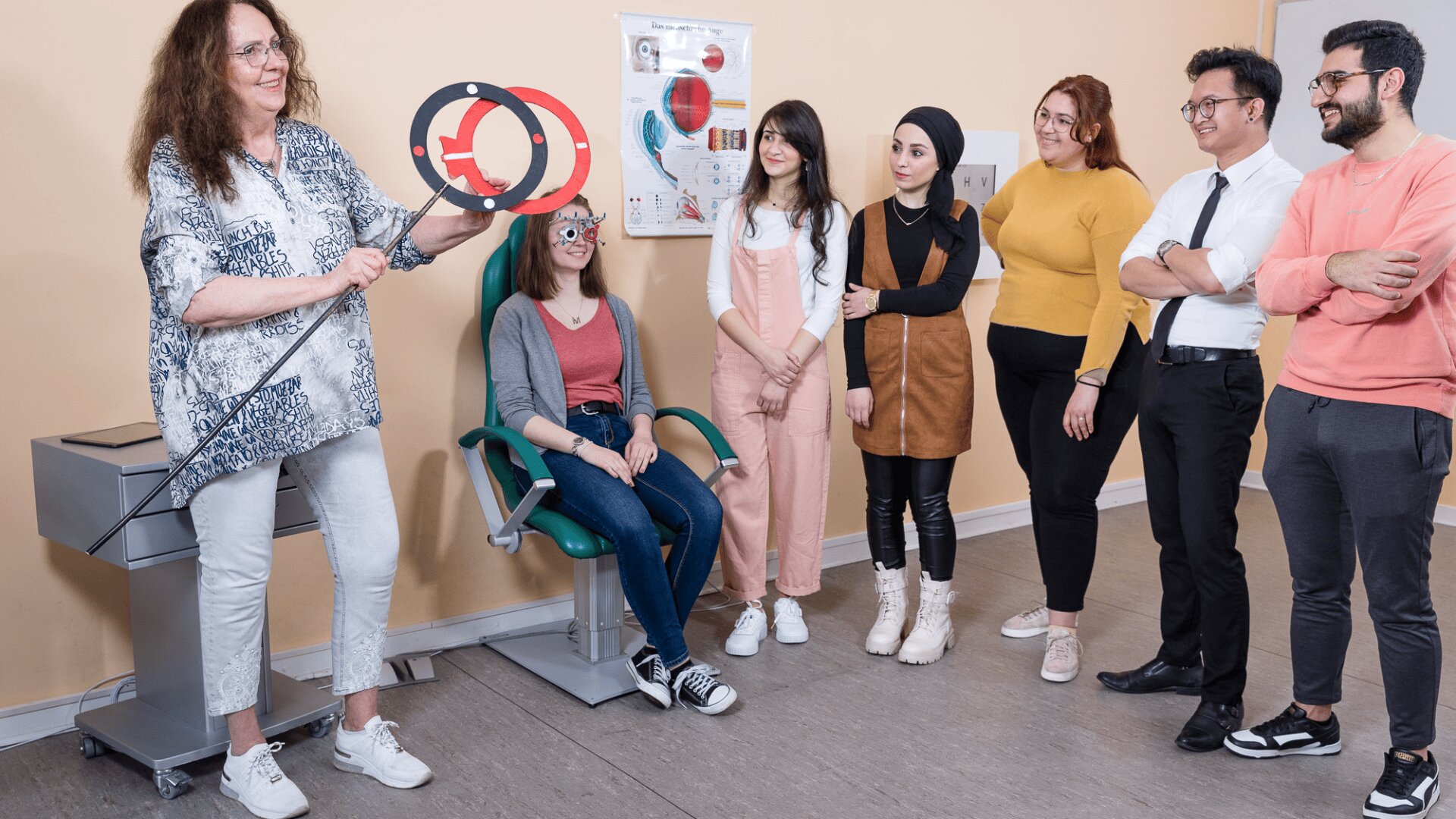 Eine Frau hält ein Sehtestgerät in der Hand und demonstriert es einer Gruppe von fünf Schülern. Ein Schüler, der auf einem grünen Stuhl sitzt, trägt eine Sehtestbrille. Die anderen stehen in der Nähe und beobachten aufmerksam die Situation in einem Klassenzimmer.