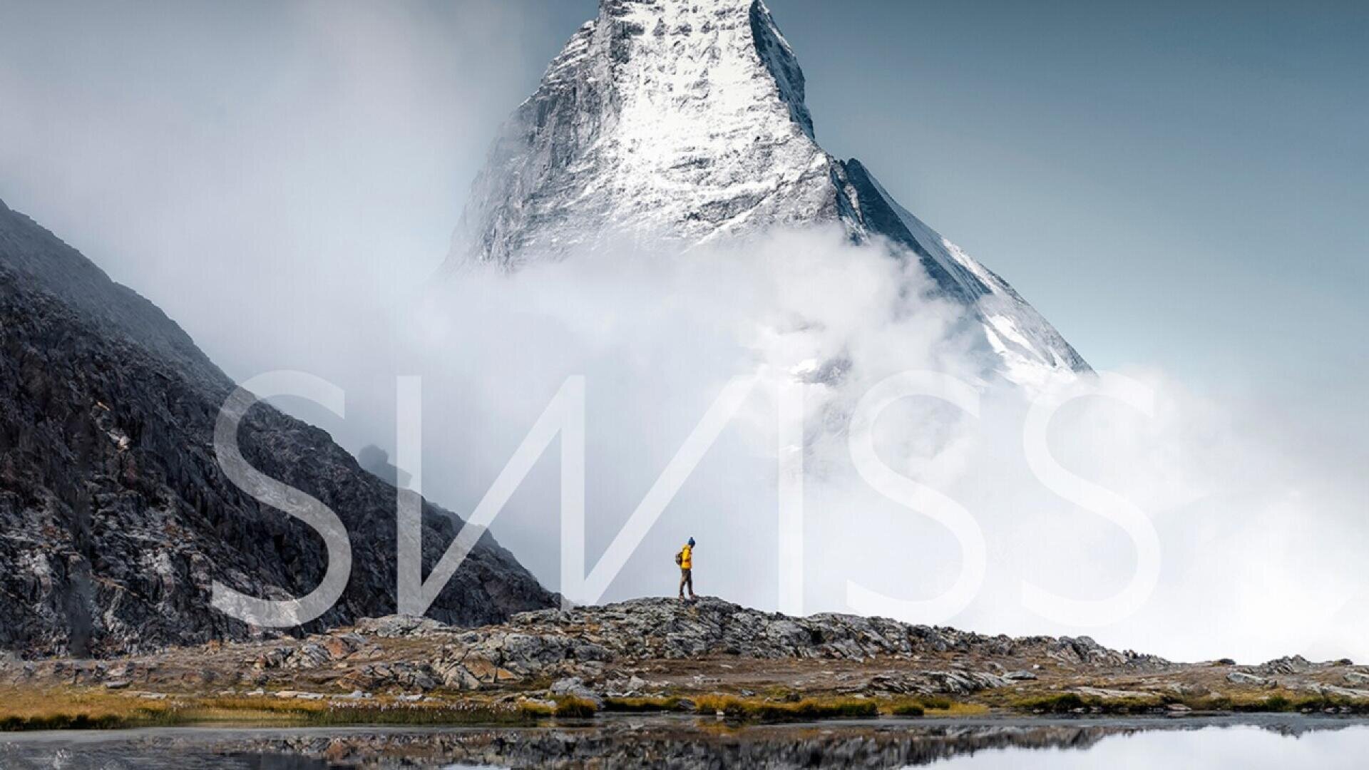 A person with a yellow backpack stands on rocky terrain, facing a snow-covered mountain peak surrounded by clouds, with the word SWISS overlaid on the image.