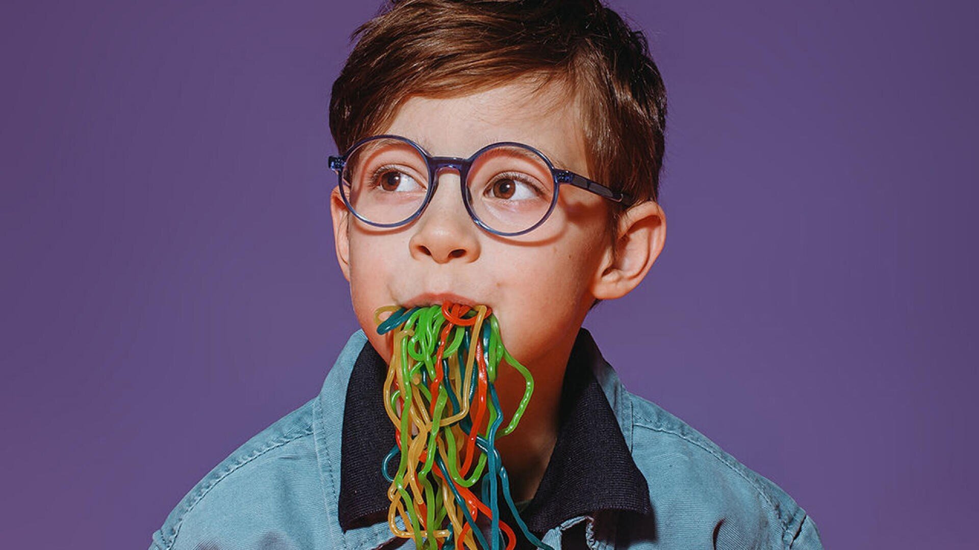 Ein kleiner Junge mit Brille und braunem Haar trägt eine blaue Jacke und vor einem violetten Hintergrund hängen bunte Bonbonketten aus seinem Mund.