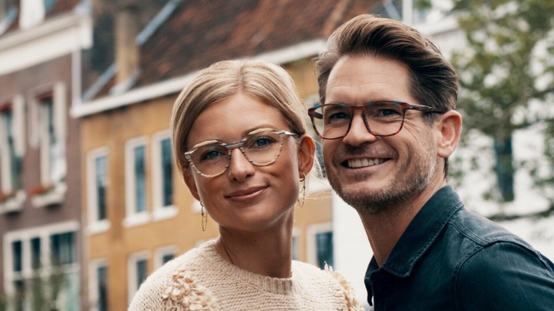A smiling woman and man wearing glasses stand close together outdoors, with blurred buildings and greenery in the background.