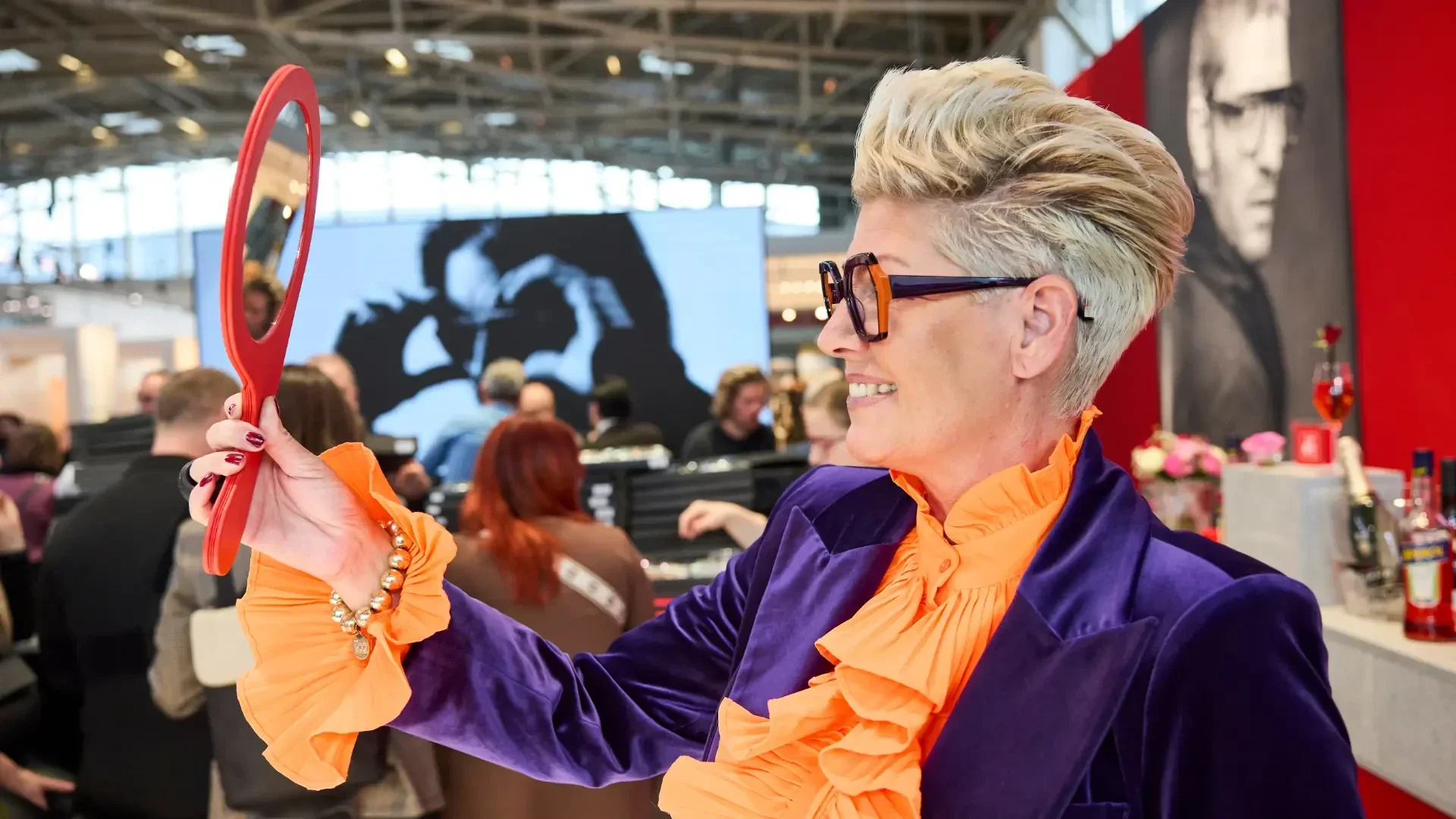 A stylish person with short blonde hair, glasses, and a purple velvet suit with orange ruffles smiles while looking into a red hand mirror at a busy indoor event.