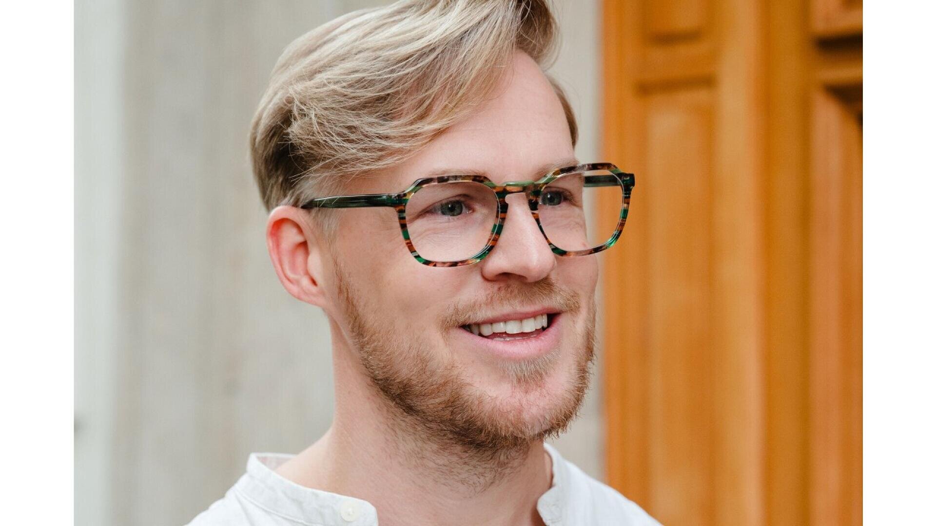 A smiling man with blond hair and a short beard wears large, round tortoiseshell glasses and a white shirt, standing in front of a wooden door and light-colored wall.