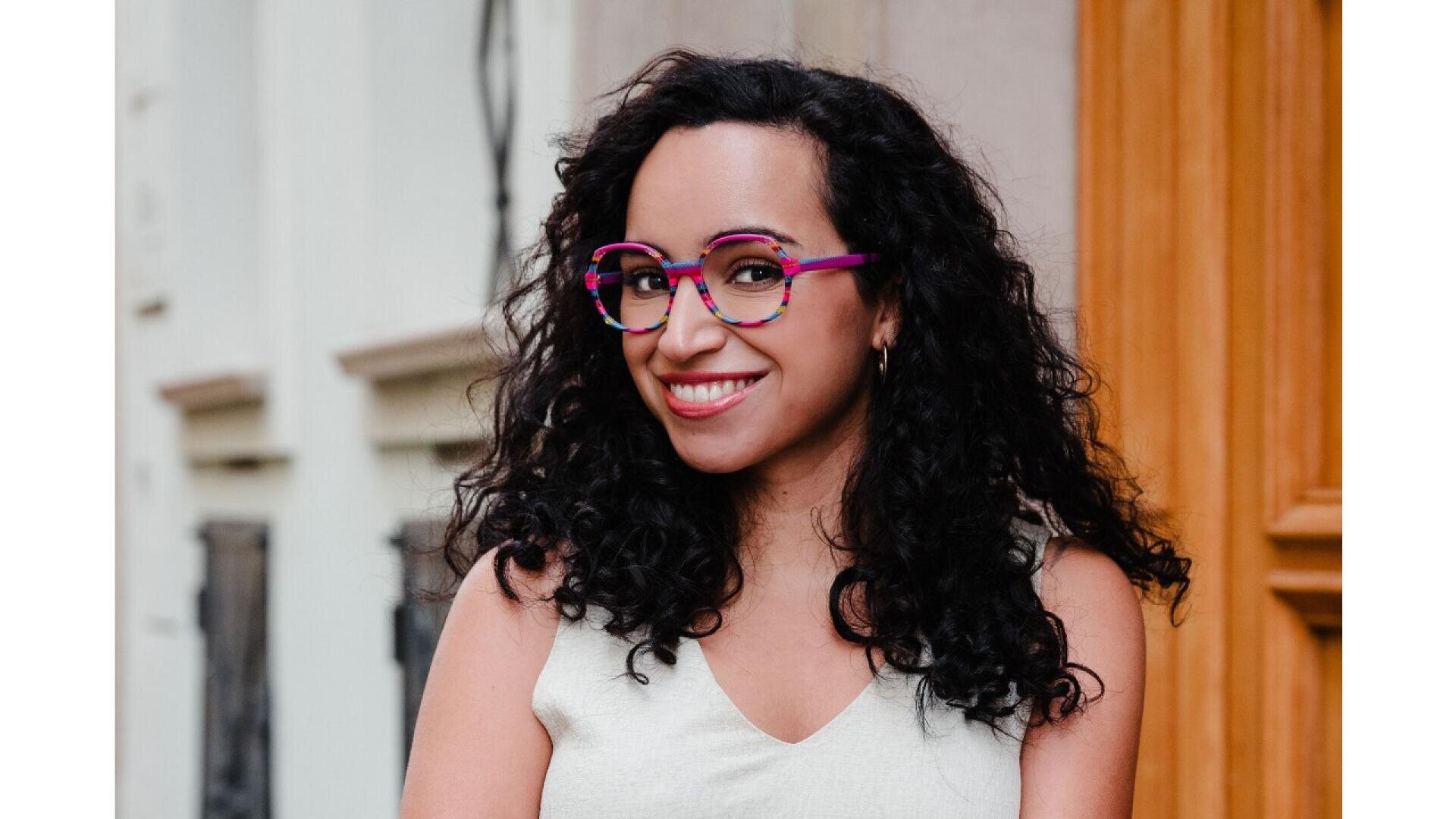 A woman with curly black hair and bright pink glasses smiles while standing outside in front of a tan building. She is wearing a sleeveless off-white top.