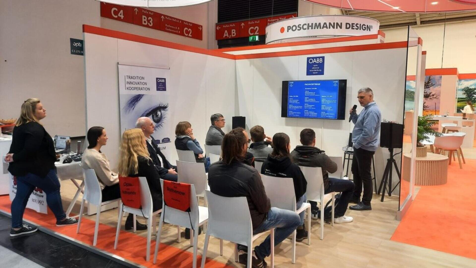 A group of people sit on white chairs listening to a man giving a presentation in a booth at a trade show. A large screen and banners are displayed, and the booth is labeled POSCHMANN DESIGN.