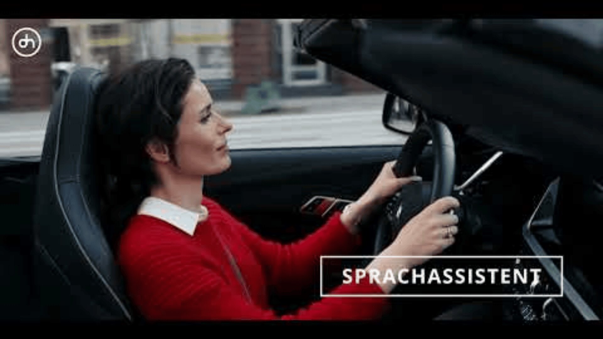 A woman in a red sweater drives a convertible car with the roof down. She is smiling and looking ahead. The word “Sprachassistent” appears on the image.