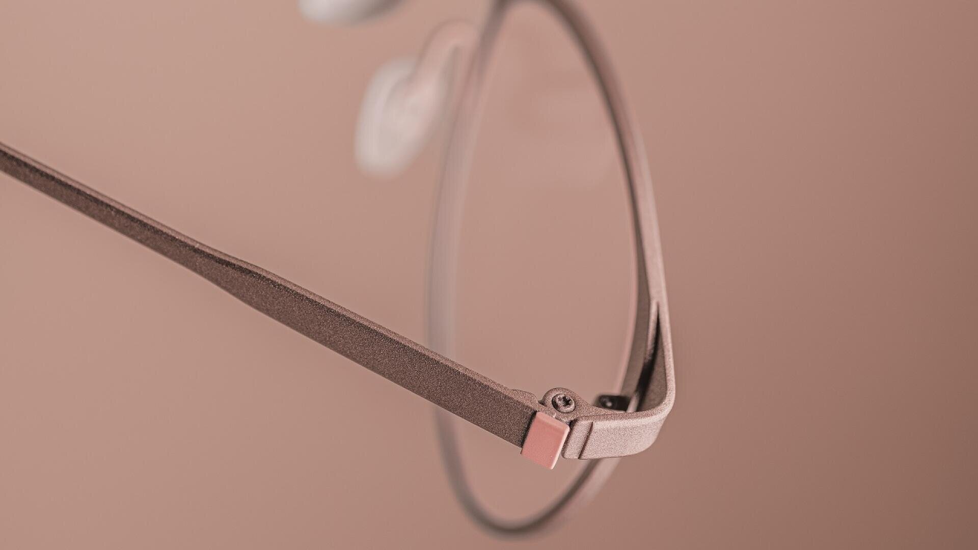 Close-up of a pair of eyeglasses with a thin, matte pinkish-brown frame, set against a matching soft pink background. Only part of the lens and the arm of the glasses are clearly visible.