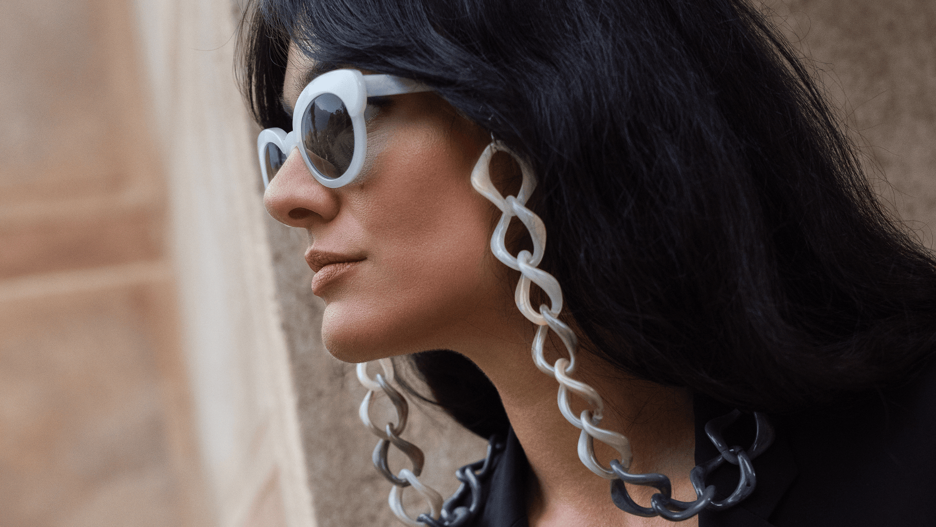A woman with long dark hair wears oversized white sunglasses with thick, chunky chain attachments and a black outfit, standing against a neutral-colored wall.