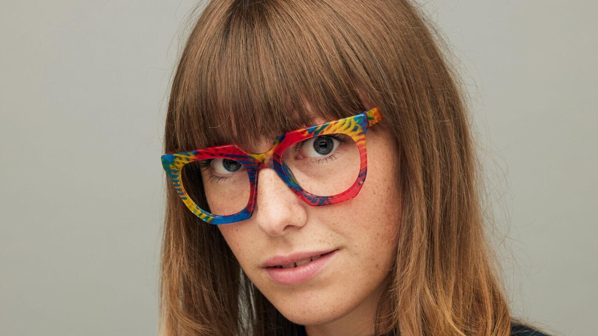 A woman with long brown hair and bangs wears oversized, colorful, patterned glasses and looks at the camera against a plain gray background.