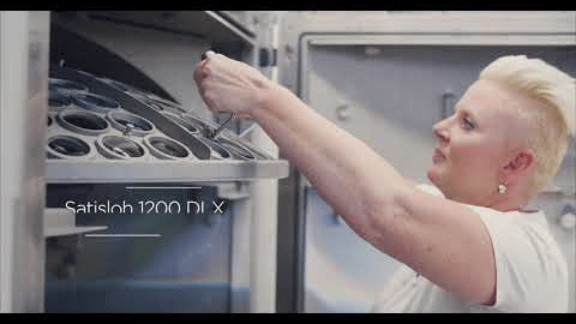 A person with short blond hair adjusts equipment inside a large industrial machine labeled “Satisloh 1200 DLX,” using a tool in a bright, clean workspace.