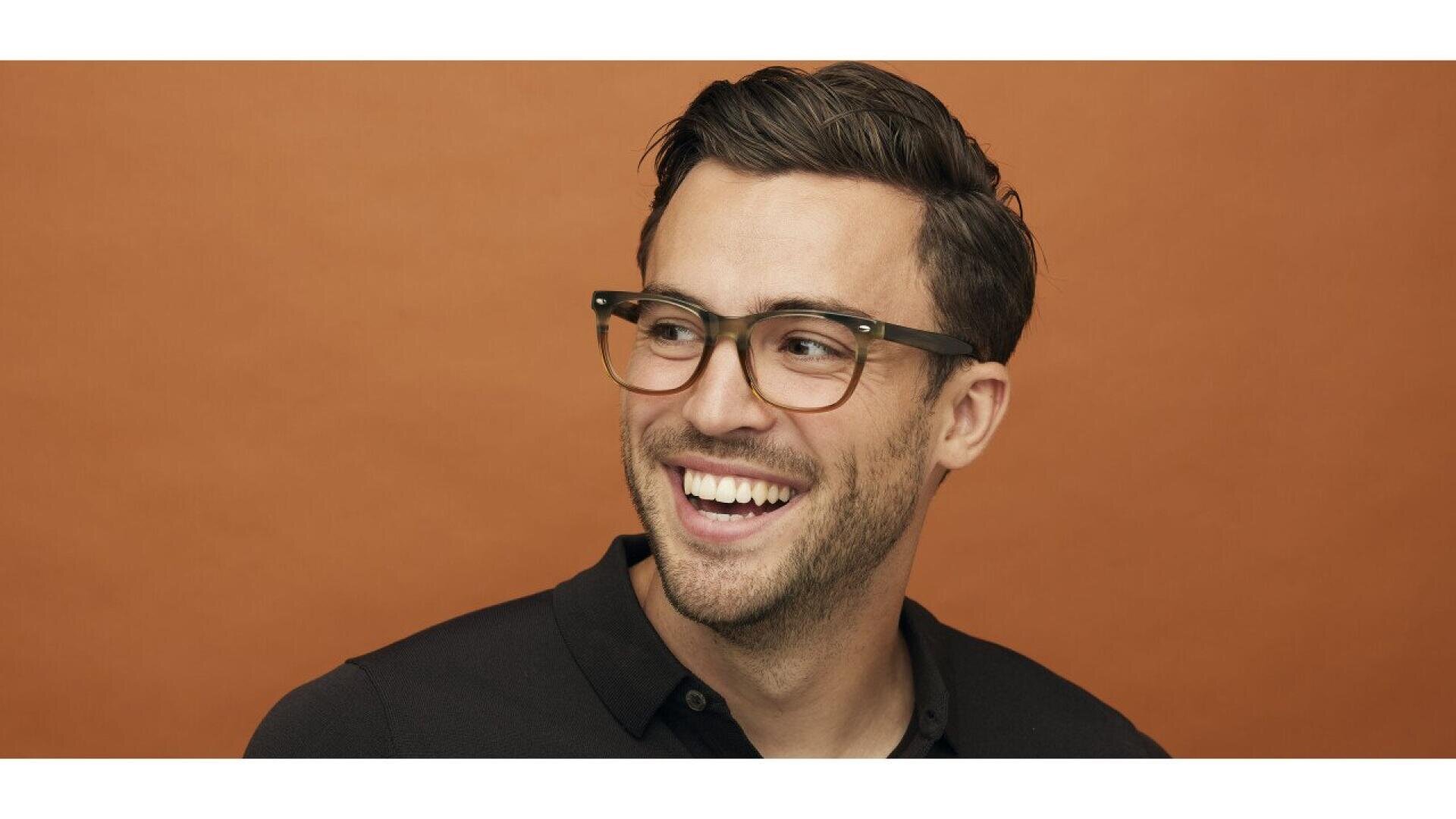 A man with short brown hair and a trimmed beard, wearing black-rimmed glasses and a black polo shirt, smiles while looking to his left against an orange background.