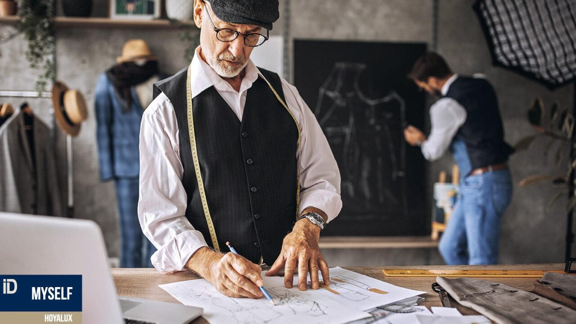 An older man in a vest and cap sketches fashion designs at a desk with sewing patterns, while another man works at a chalkboard with suit patterns in a stylish tailor’s studio.
