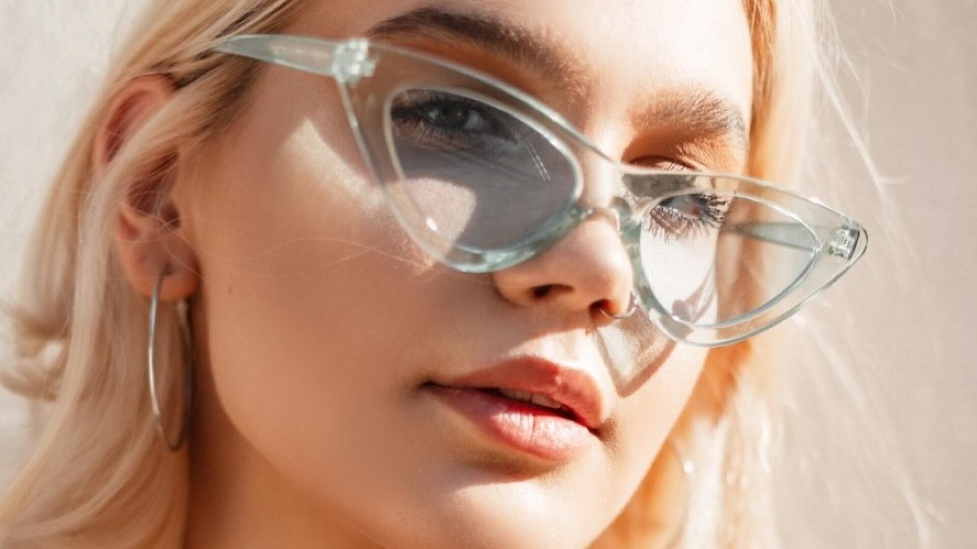 Nahaufnahme einer Frau mit hellblondem Haar, die eine klare Katzenaugenbrille und große Ohrringe trägt und bei natürlichem Sonnenlicht selbstbewusst in die Kamera schaut.