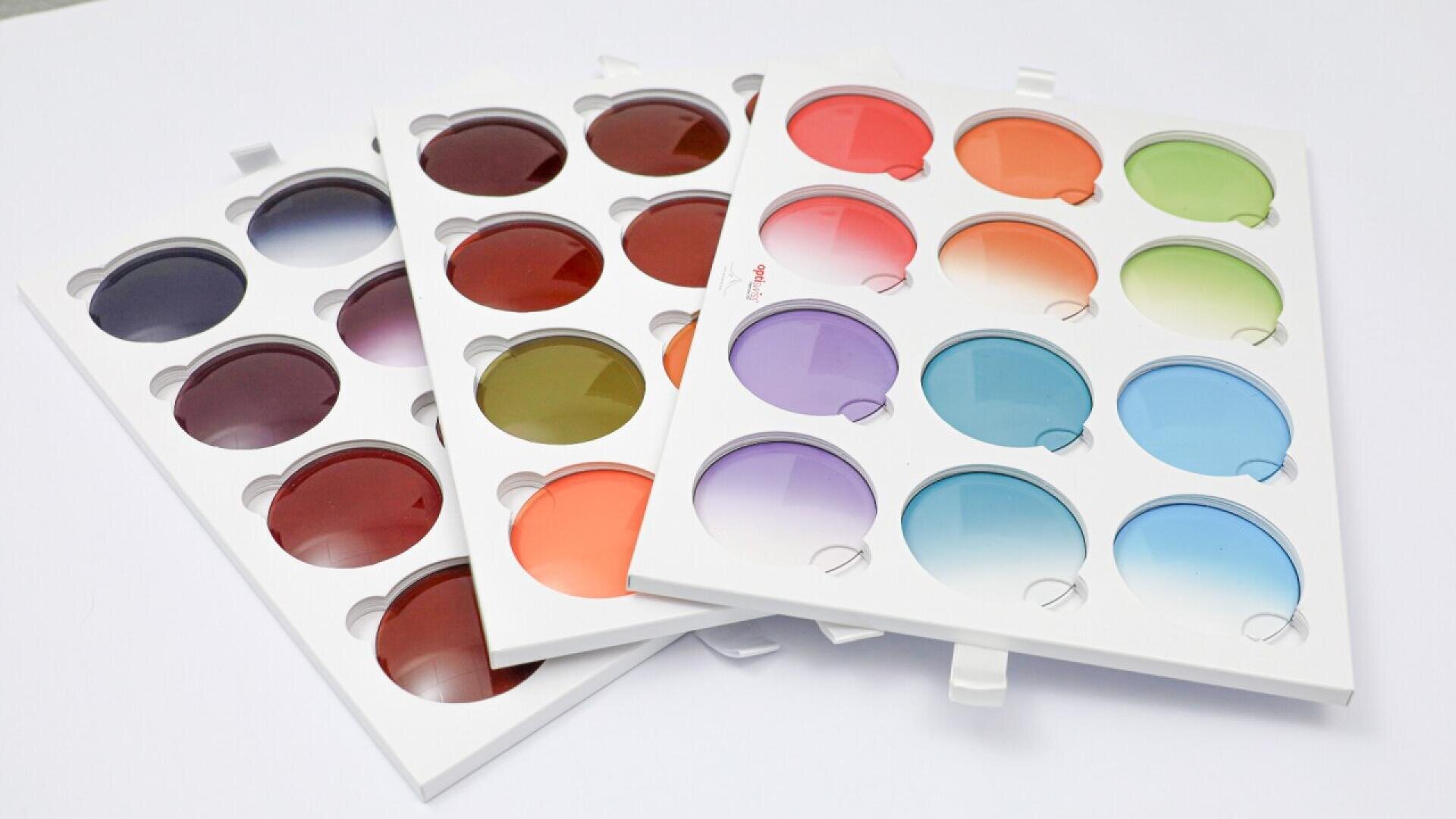 Three white trays display various round sunglasses lenses in different colors and gradients, including red, orange, purple, blue, and green, on a white background.