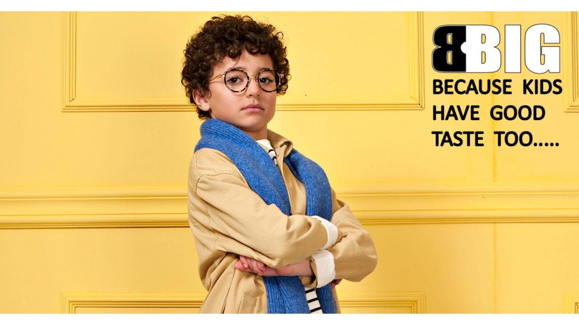 A young boy with curly hair and glasses stands confidently with arms crossed, wearing a beige coat and blue scarf, in front of a yellow wall. Text reads: BIG Because kids have good taste too.....