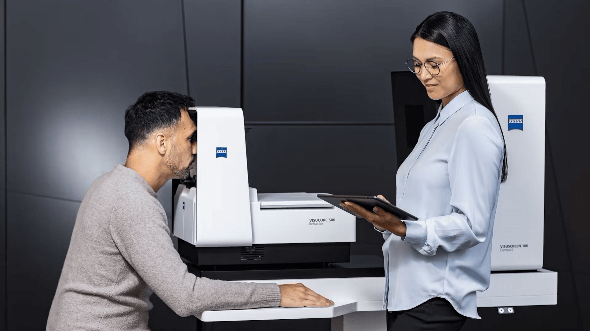 A man undergoes an eye exam using a diagnostic machine while a woman in glasses and a light blue blouse stands next to him, holding a tablet and observing the procedure.