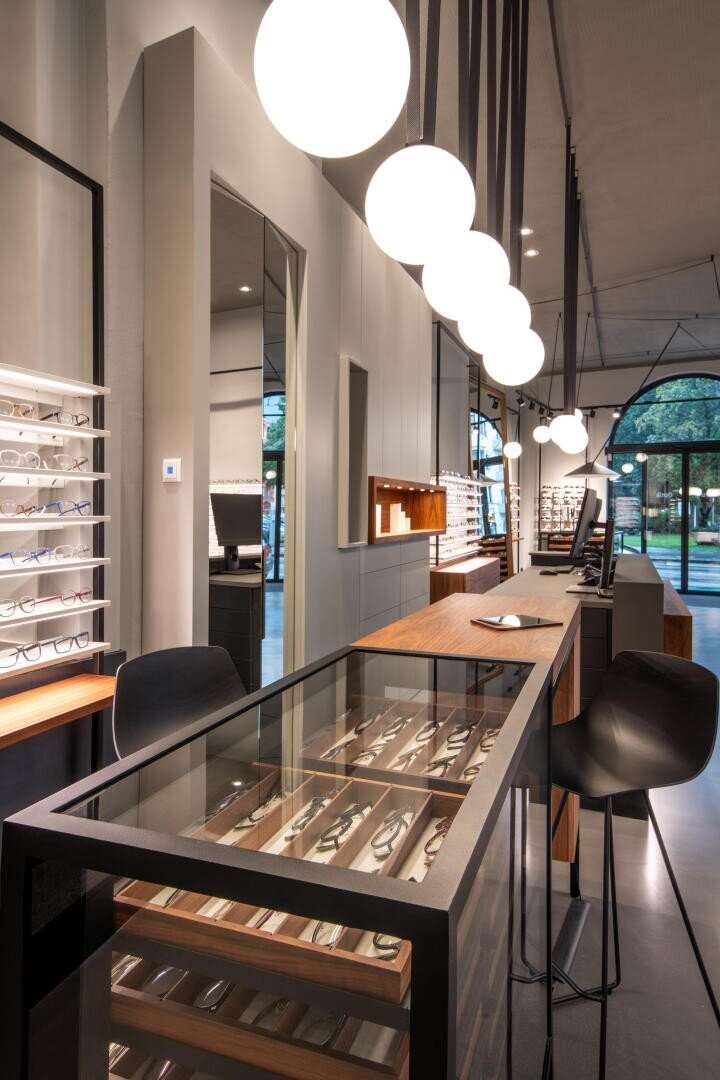 Modernes Brillengeschäft mit Glasvitrinen für Brillen, schlichten Holztheken, schwarzen Stühlen und runden, von der Decke hängenden Pendelleuchten. Große Fenster lassen natürliches Licht herein.