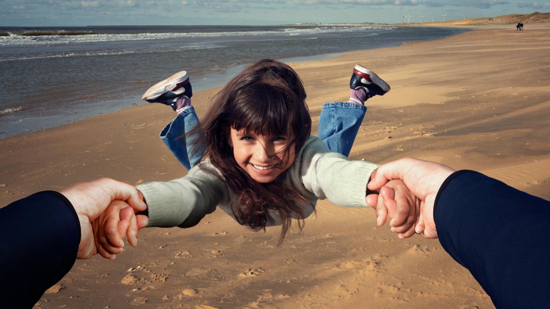 A smiling young girl is held by her hands and swung above the sandy beach by an unseen adult, with the ocean and blue sky in the background.