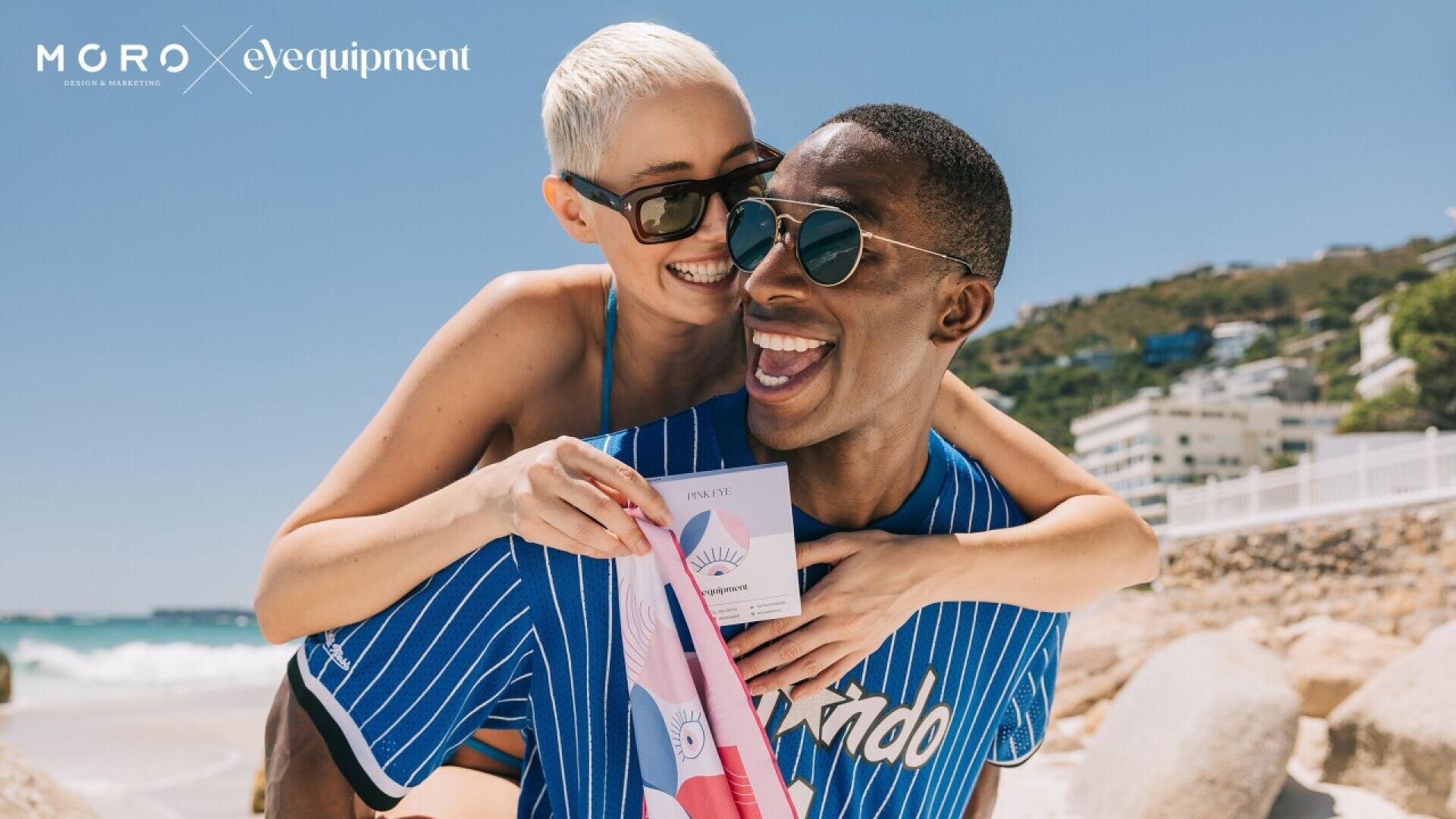 A cheerful couple wearing sunglasses smiles and poses playfully on a sunny beach; the woman hugs the man from behind as he holds a colorful shirt and a box labeled “Privé Revaux.” Logos read “Moro” and “Eyequipment.”.