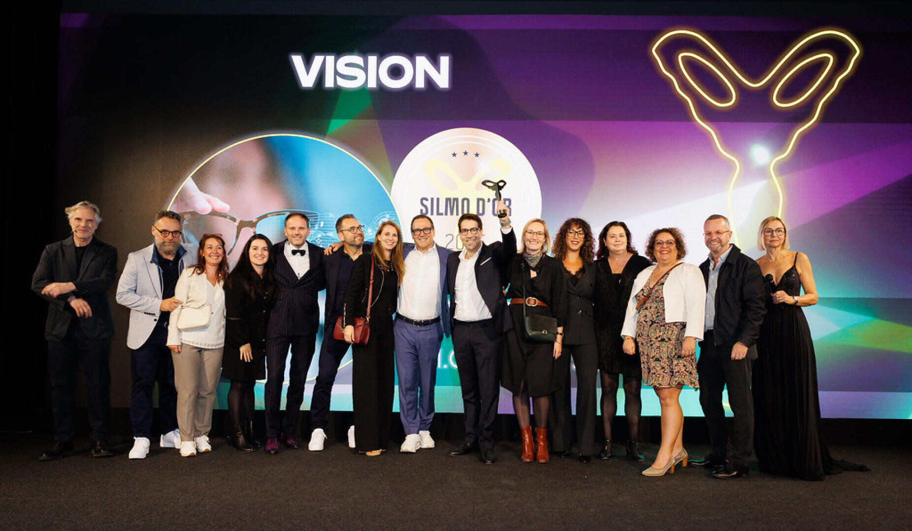 A group of 16 people stand on a stage under the word "VISION" and smile for a photo. One person in the middle holds up a trophy. Colorful graphics and a glowing outline can be seen on the screen behind them.