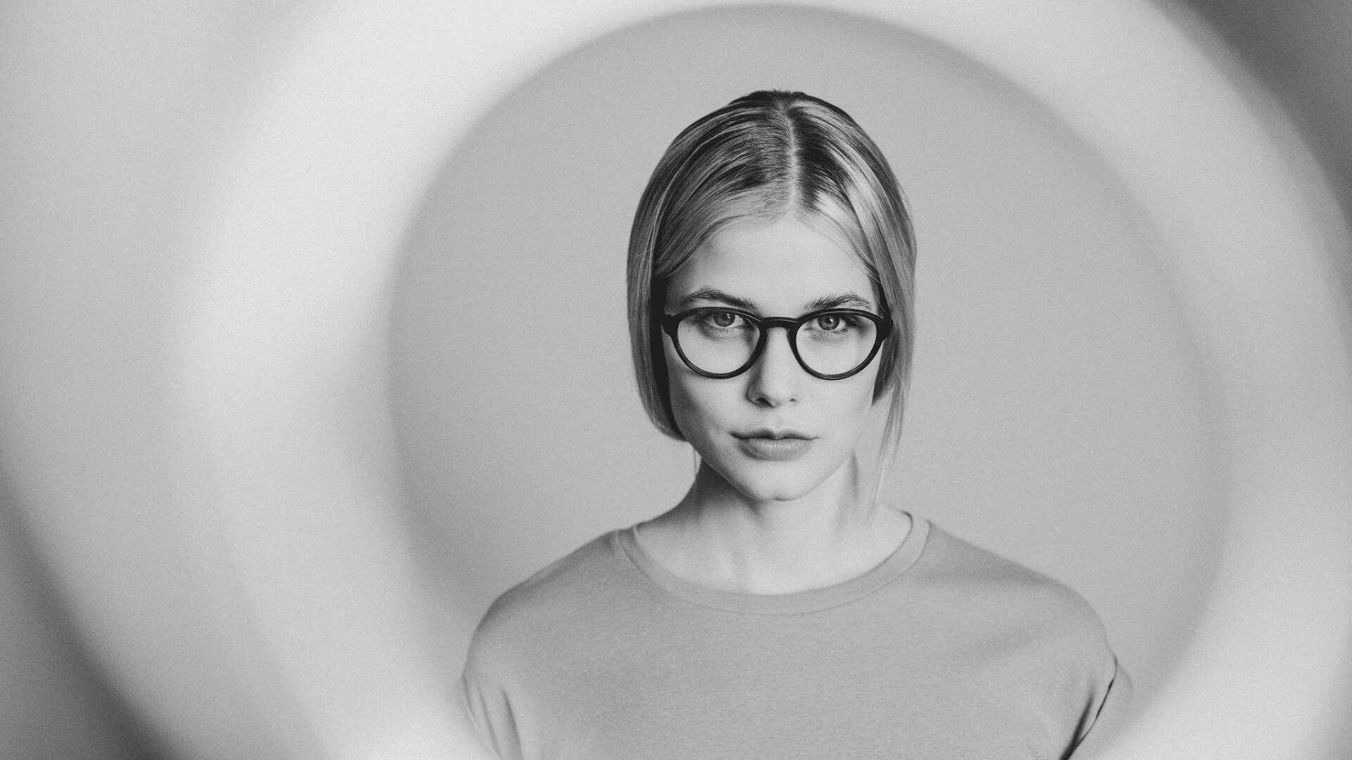 Schwarzweißfoto einer Frau mit glattem, schulterlangem Haar und runder Brille, zentriert in einem kreisförmigen Lichtring, die mit neutralem Gesichtsausdruck direkt in die Kamera blickt.